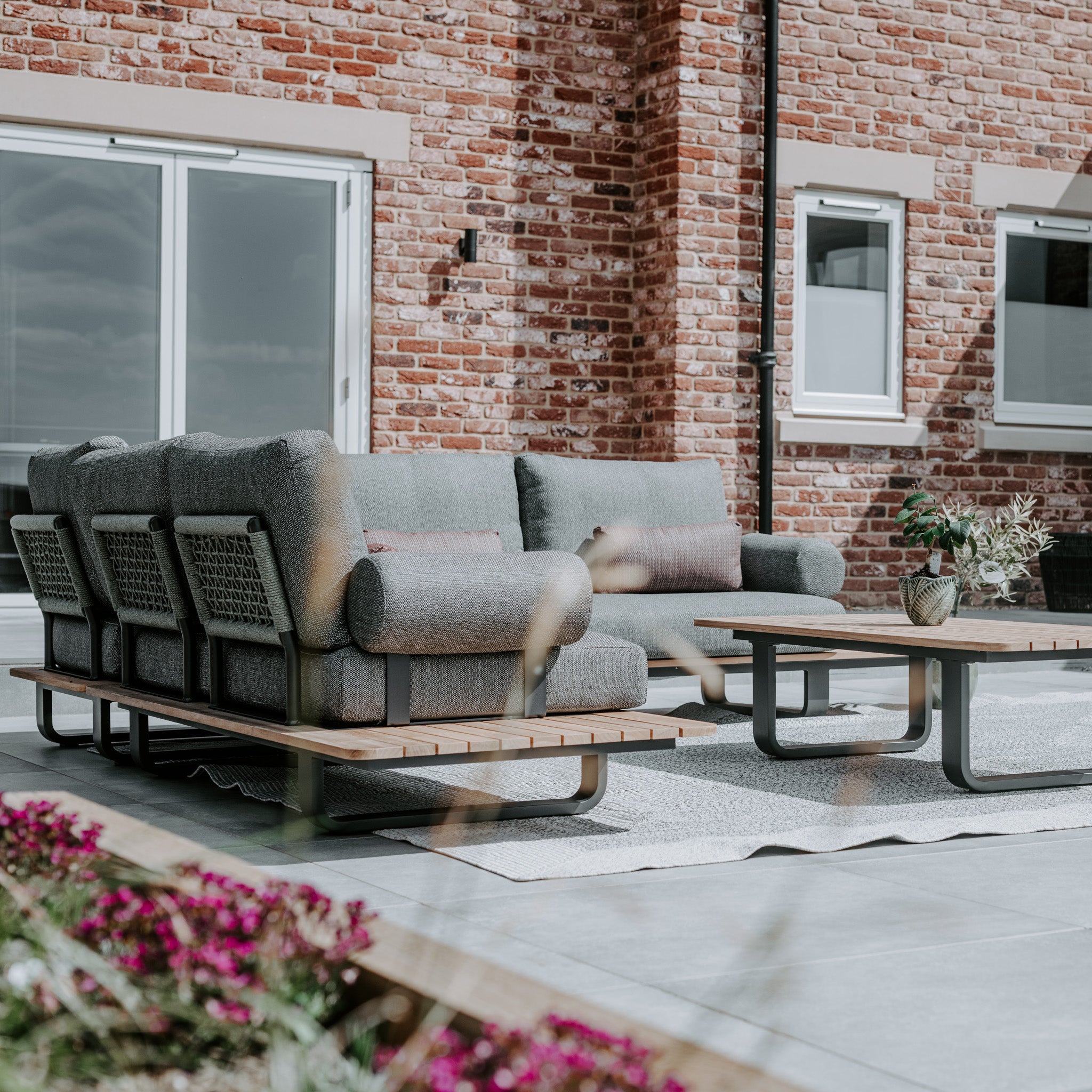 Modern outdoor patio with a Bay Corner Group Set featuring 4 waist pillows, an olive green teak table, and potted plants against a red brick wall backdrop.