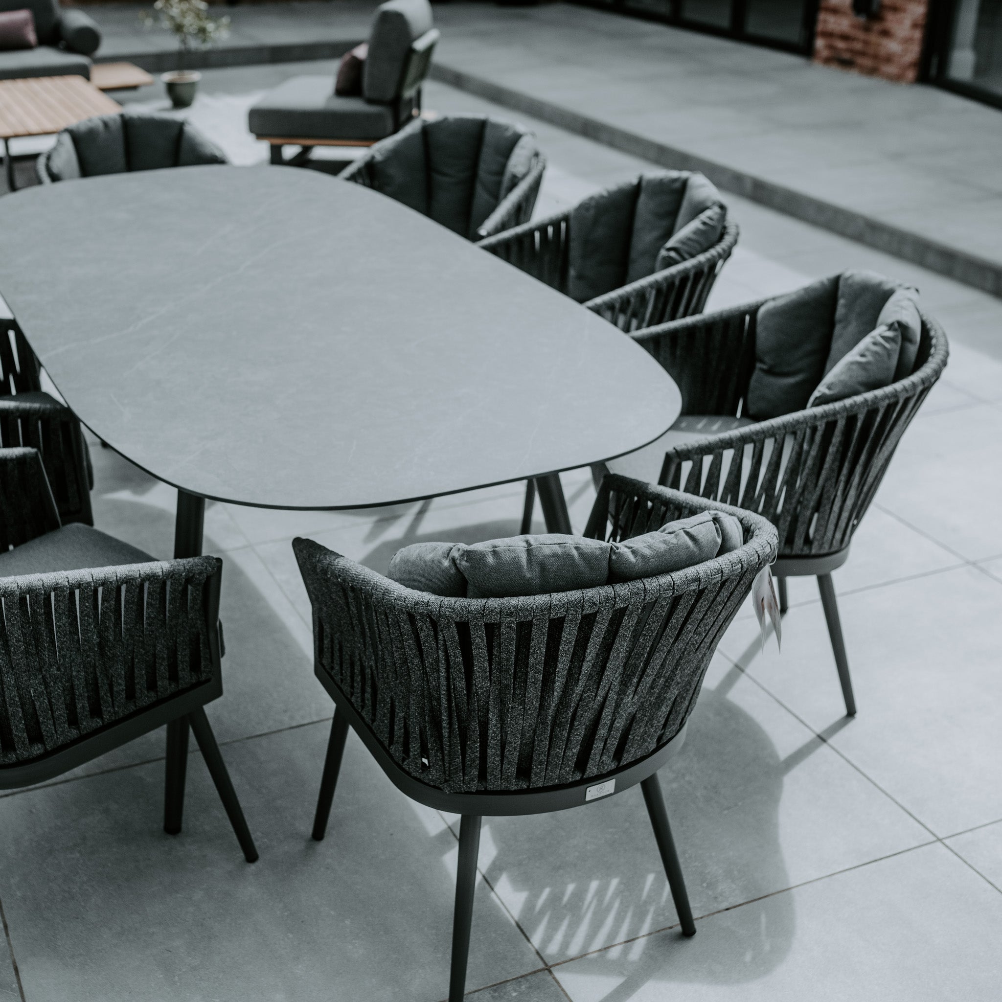 Outdoor patio with a Monterrey 8 Seat Rope Dining Set featuring an oval ceramic table in grey, surrounded by six cushioned woven chairs on a concrete tile floor.