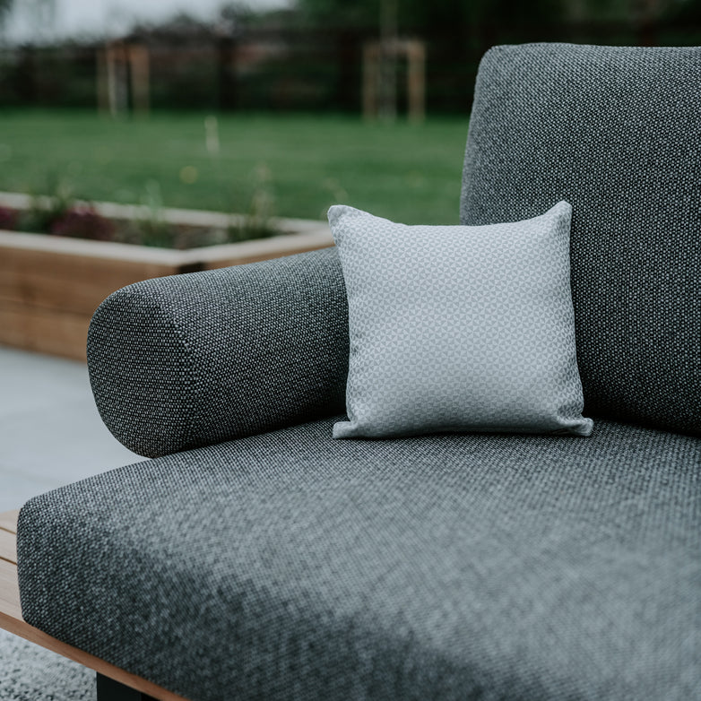 A gray outdoor sofa with a light gray pillow on a patio, featuring green lawn and garden planter in the background. Enhance your space with an Acrisol Helix Gris Claro Small Scatter Cushion - 25cm x 25cm for ultimate garden personalization.