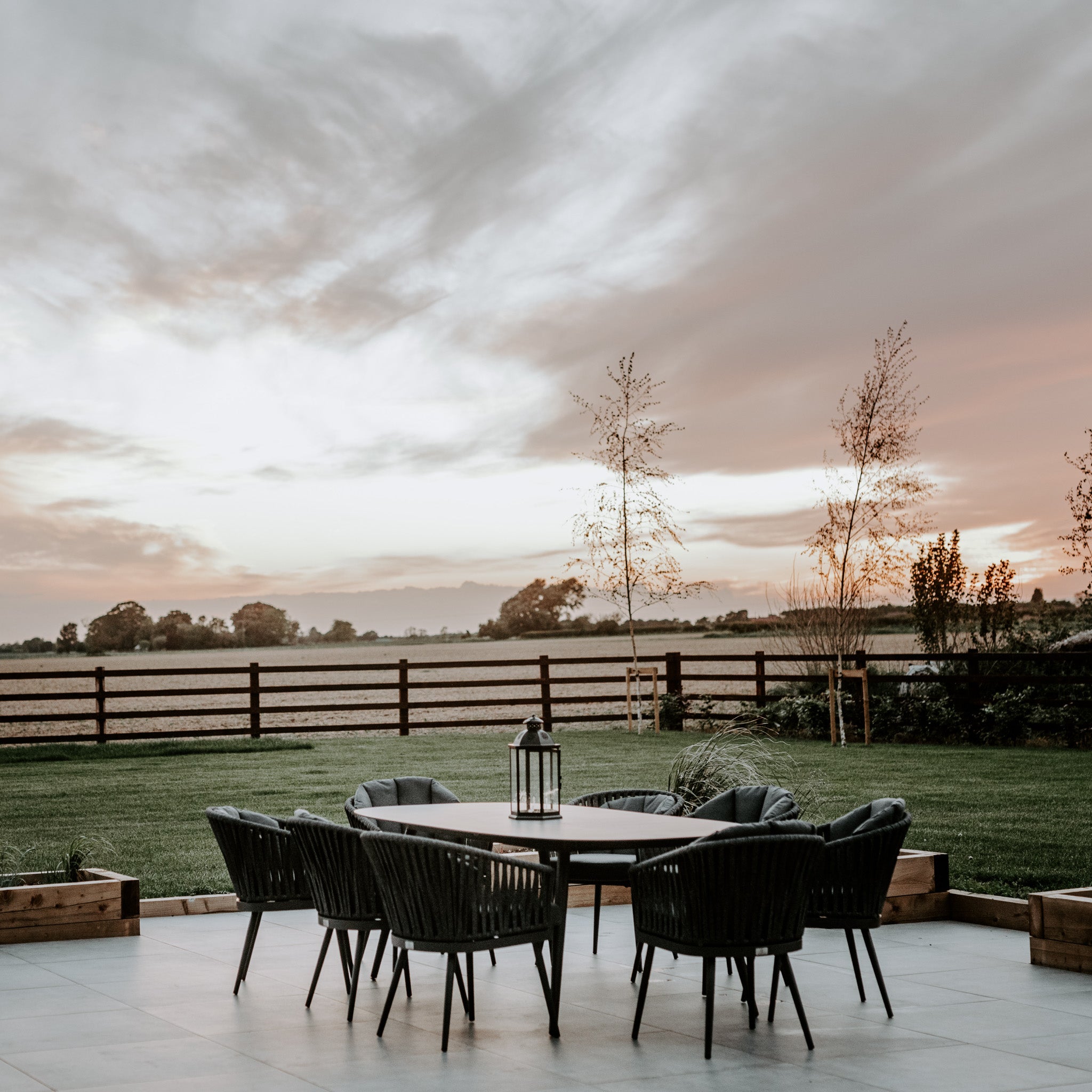 Outdoor patio with a Monterrey 8 Seat Rope Dining Set with an Oval Ceramic Table in Grey and six black chairs, facing a field under a cloudy sky during sunset.