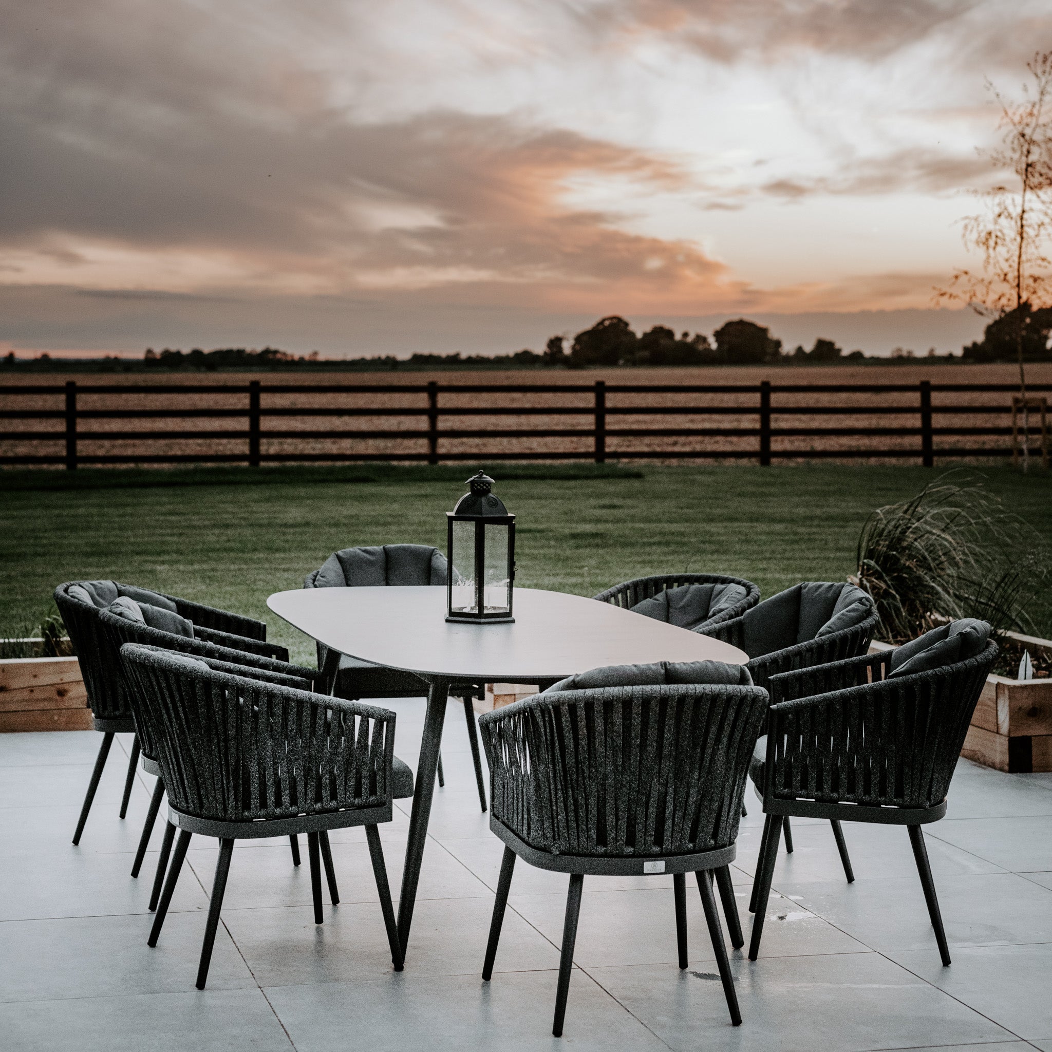 Monterrey 8 Seat Rope Dining Set with Oval Ceramic Table in Grey, featuring a lantern centerpiece and offering a perfect view of the sunset over a field.