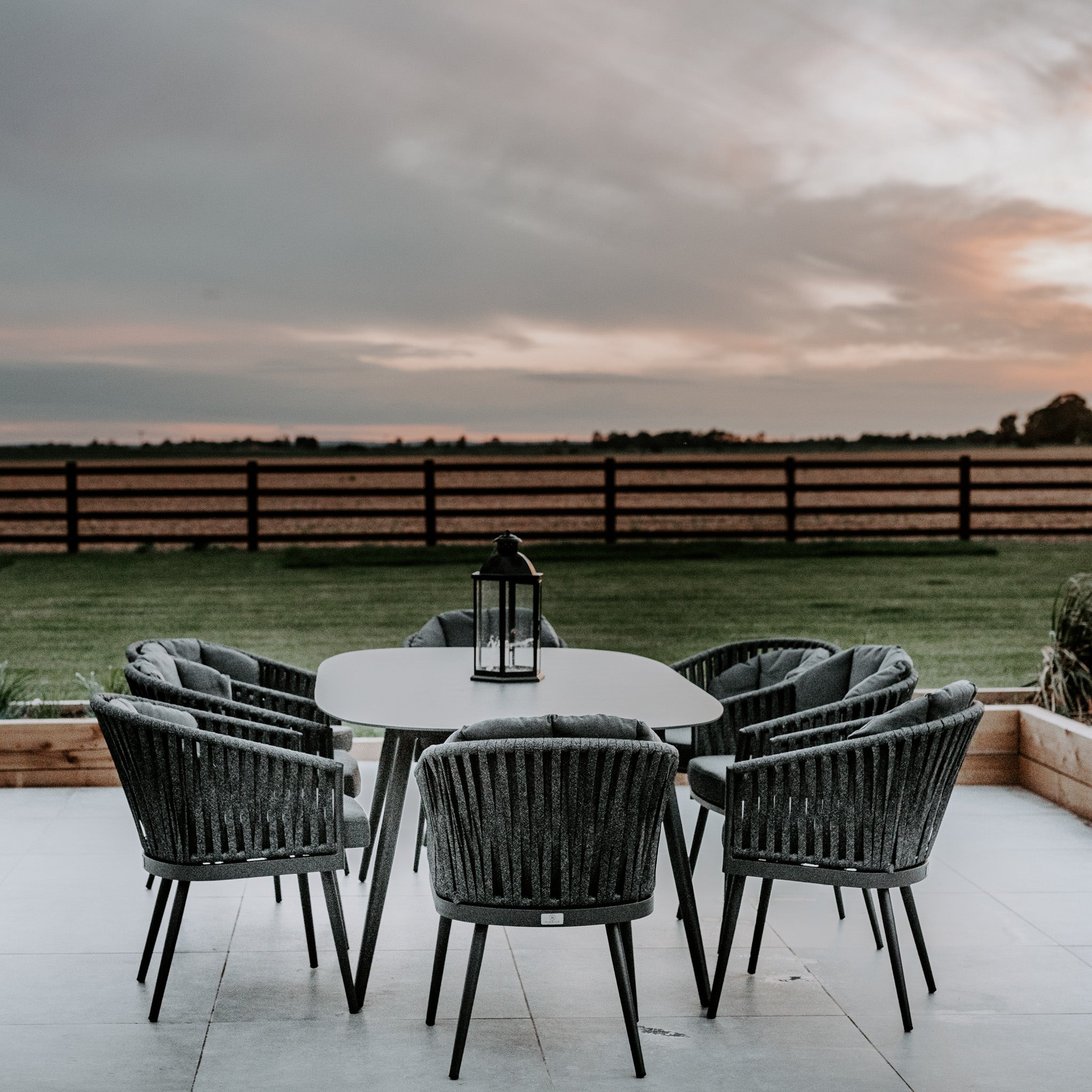 The outdoor dining area showcases a Monterrey 8 Seat Rope Dining Set with an Oval Ceramic Table in Grey, featuring six grey rope dining chairs arranged around the elegant table. A lantern serves as the centerpiece, set against a backdrop of a field enclosed by a fence at sunset.