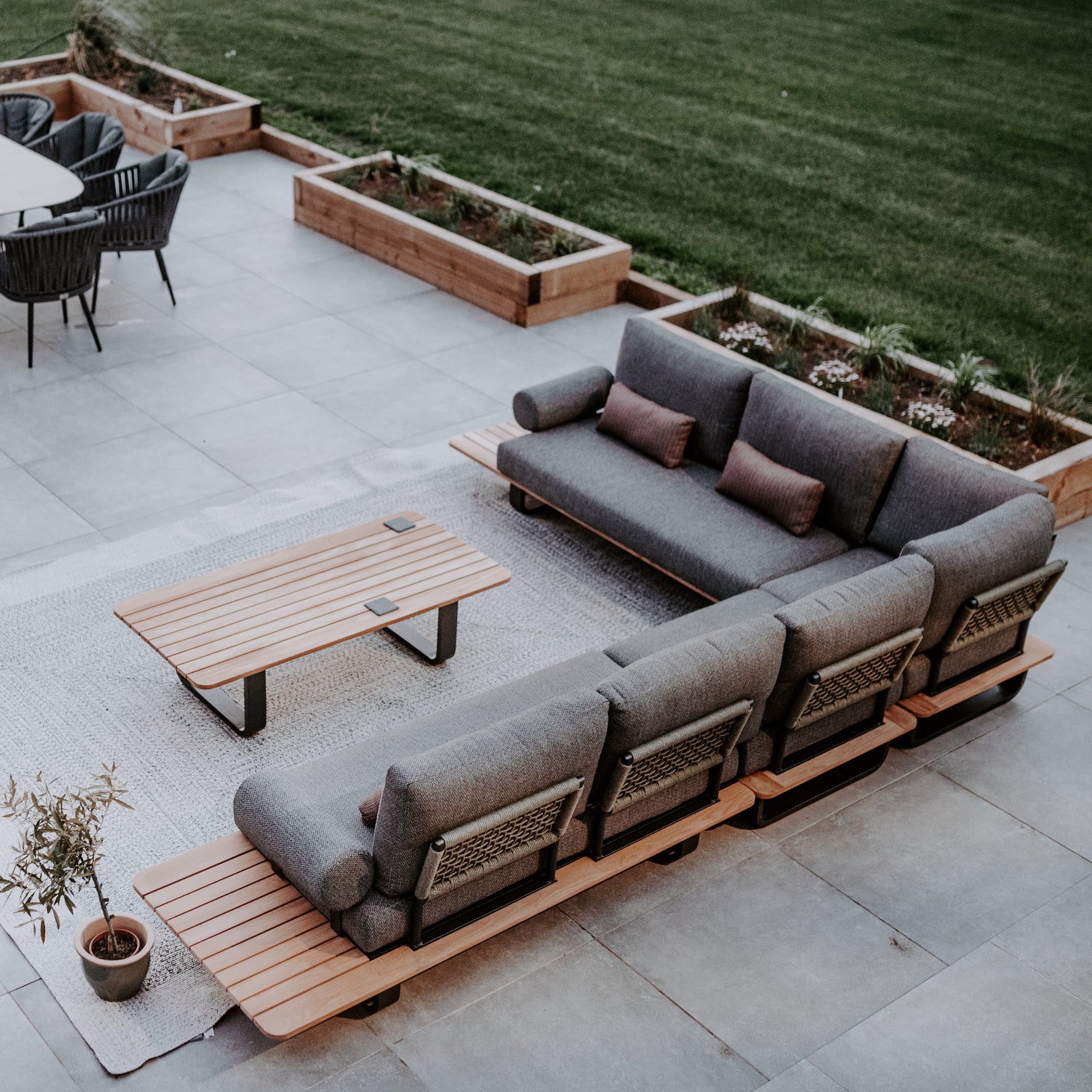 Modern patio featuring a Bay Large Corner Group Set with 4 Waist Pillows in Olive Green and a teak table, potted plant, surrounding garden beds, and outdoor dining area.