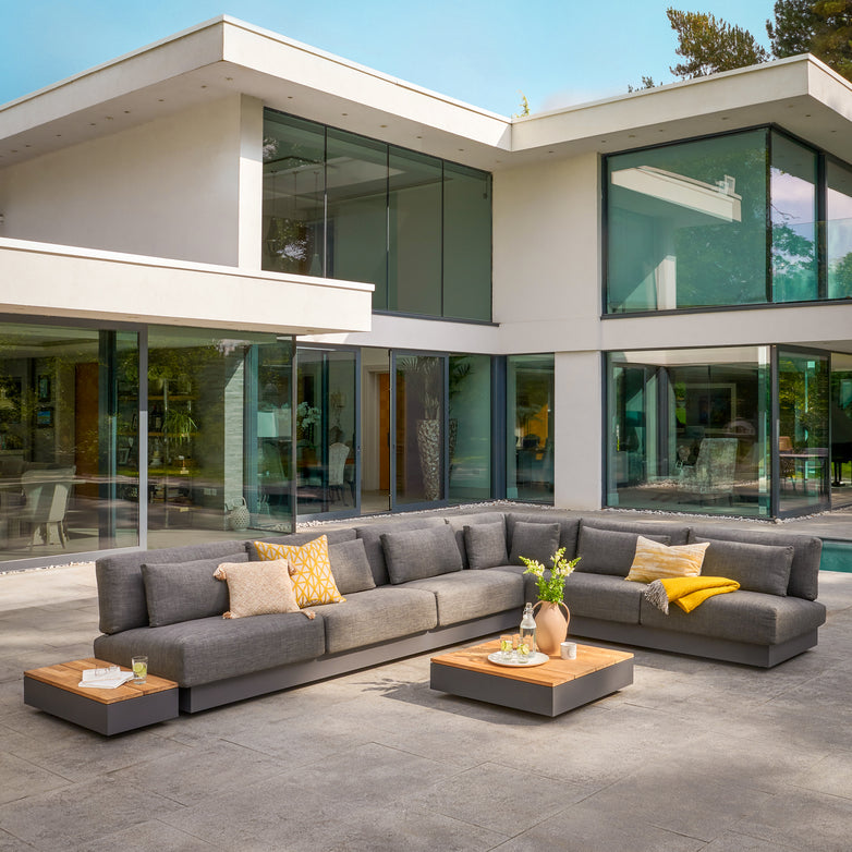 Modern outdoor patio showcasing the Ibiza Corner Group Set with Teak Tables in Anthracite, styled with yellow pillows and low teak-topped tables, elegantly placed before a striking glass house.