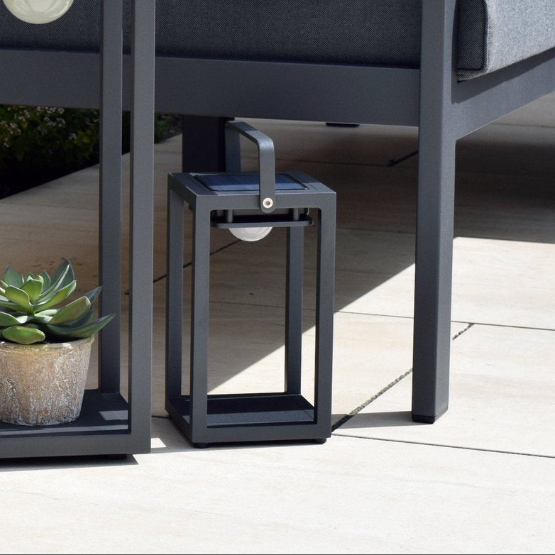 Two Luxor Small Table Outdoor and Indoor Solar Lanterns in charcoal, one larger with a succulent plant inside, sit next to a grey outdoor sofa adorned with white and grey cushions. These weatherproof lights not only enhance the ambiance but also ensure durability outdoors.