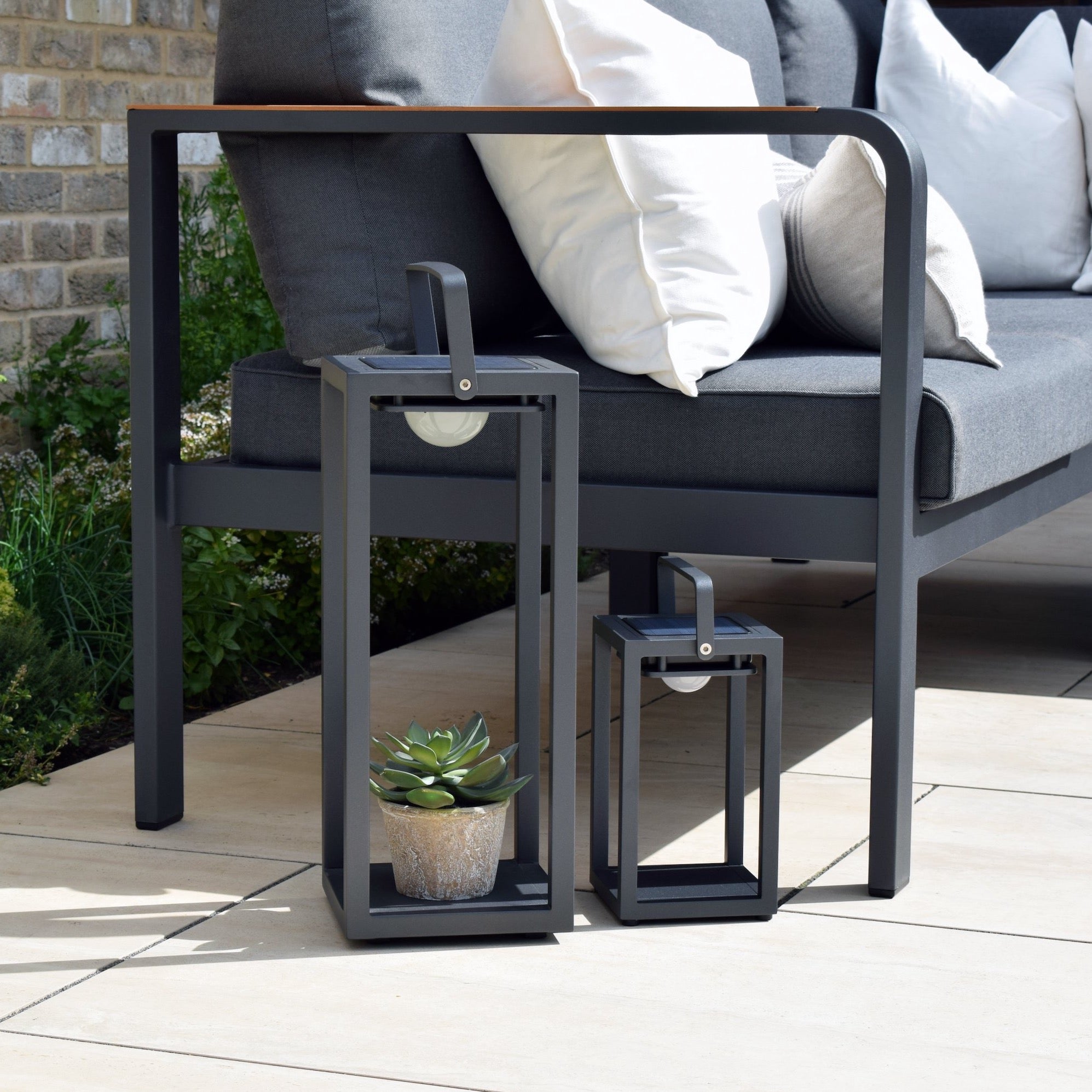 Two Luxor Large Floor Outdoor and Indoor Solar Lanterns in Charcoal, one larger than the other, adorned with a potted succulent inside and equipped with Solar lighting for a cozy evening ambiance, placed next to a modern outdoor sofa.