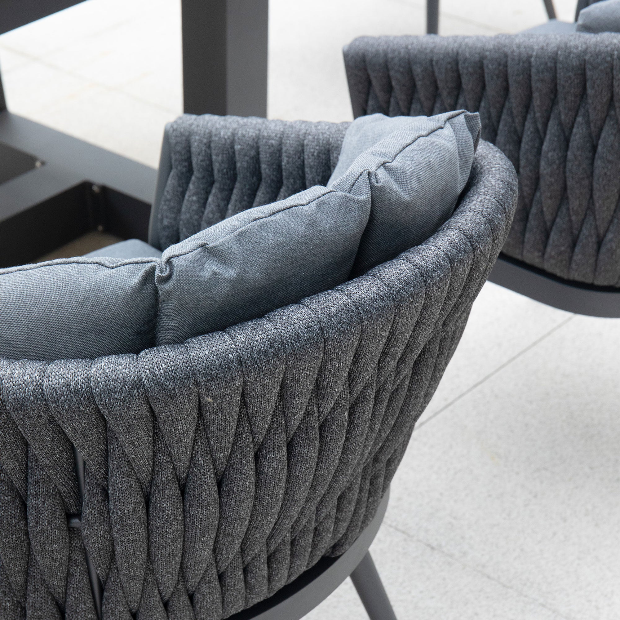 Close-up of modern grey woven fabric chairs with cushions, arranged around a table on a light-colored patio, featuring the sleek Palma 8 Seat Rope Rectangular Teak Firepit Dining Set in Grey with durable aluminium frames.