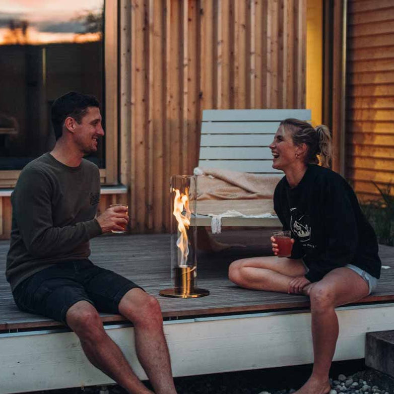 Two people are sitting on a wooden deck, smiling and holding drinks beside the warm glow of their Höfats SPIN 120 Table-Top Fire in Gold.
