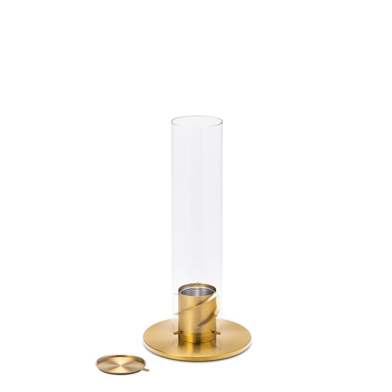 A modern glass and gold candle holder with a cylindrical shape and a removable lid is placed next to it on a white background, mirroring the elegance of the Höfats SPIN 90 Table Top Fire in Gold.