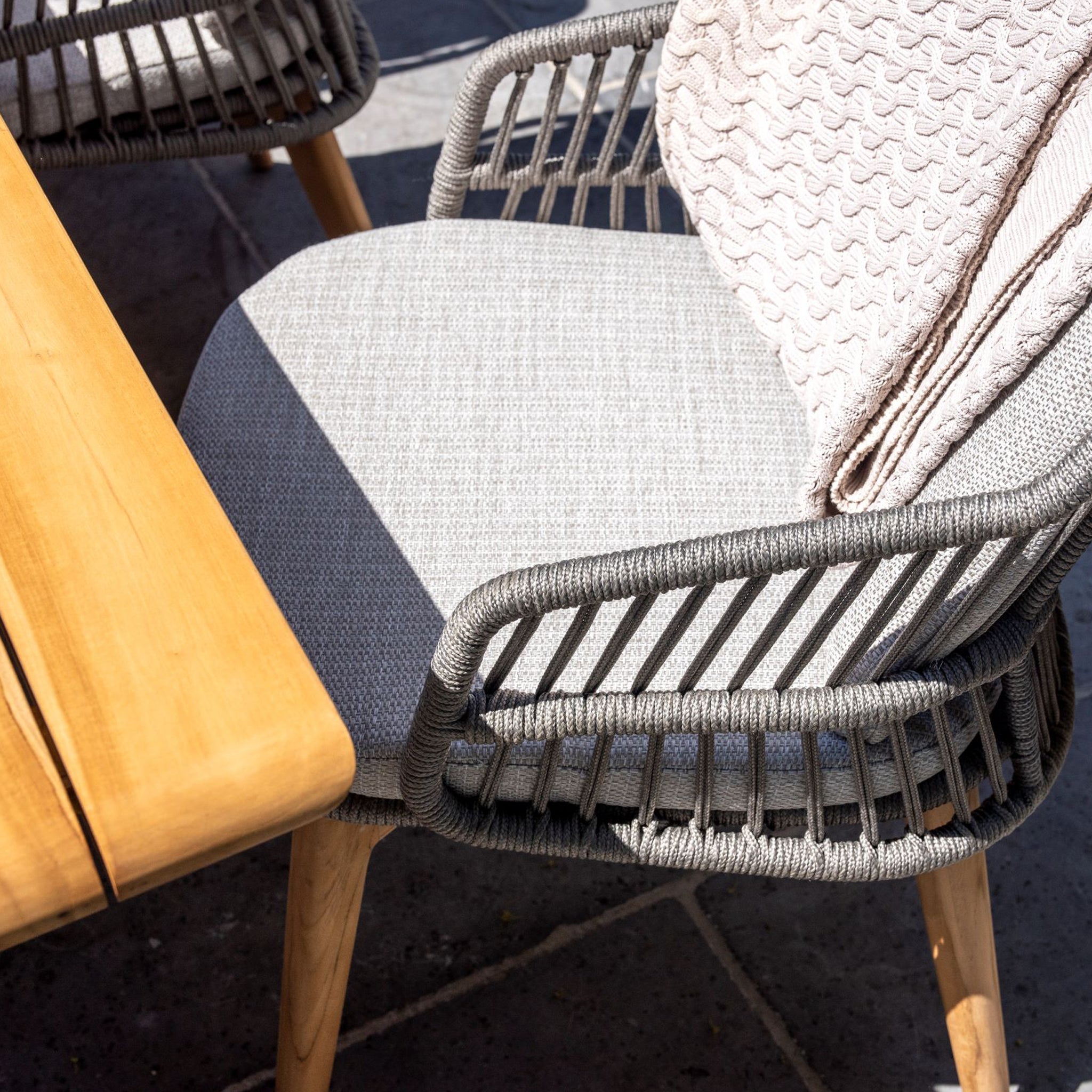 A Sempre 6 Seat Rectangular Dining Set featuring a light grey teak table includes a handmade woven outdoor chair with a gray cushion and a textured blanket draped on the back.