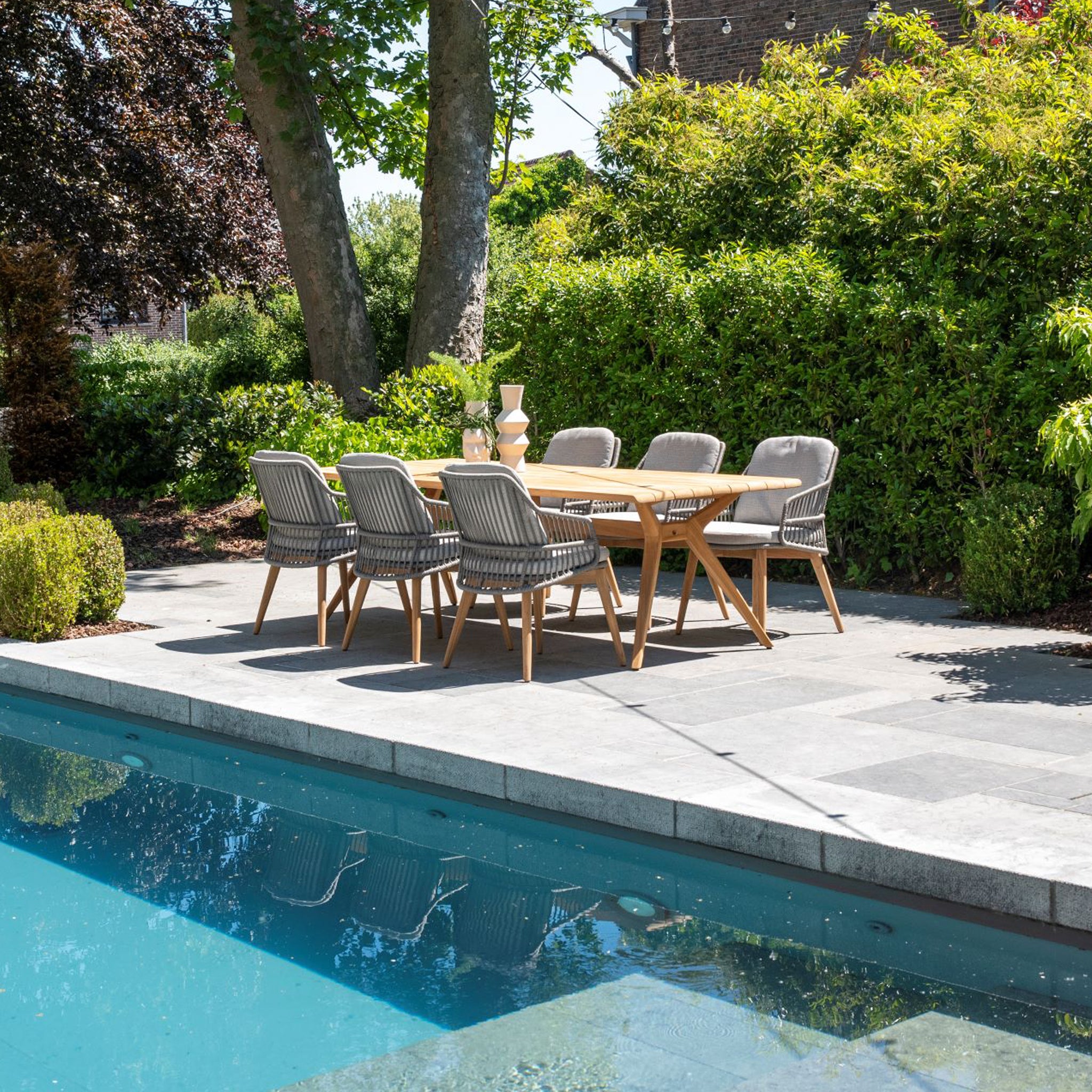 The Sempre 6 Seat Rectangular Dining Set with Teak Table in Light Grey elegantly adorns a stone patio by a pool, surrounded by lush greenery and trees in the background.