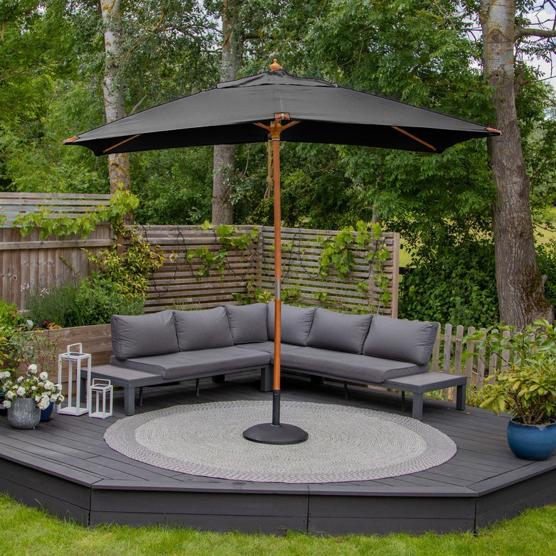 Patio with a Styx 3m x 2m Rectangular Deluxe Wooden Parasol in Grey, gray sectional sofa, and potted plants on a round rug in a lush green garden.