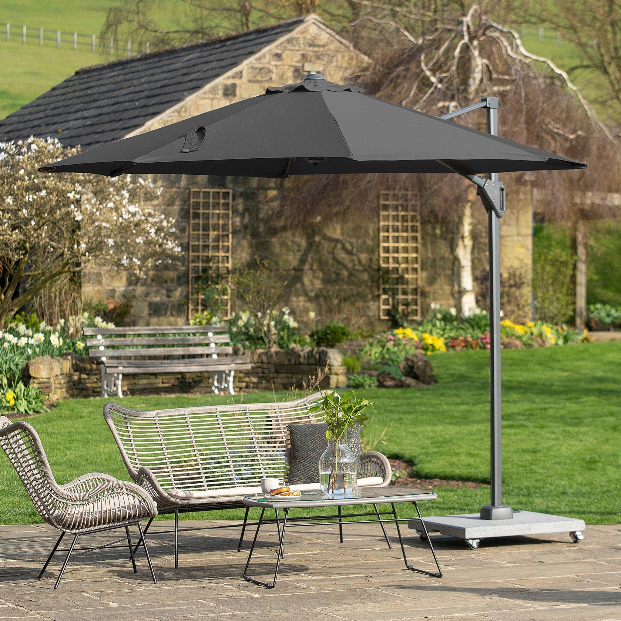 A garden with blooming flowers and a stone wall showcases a Pacific Lifestyle Voyager T1 3m round parasol in anthracite with UPF35+ protection, complemented by wicker chairs and a table.