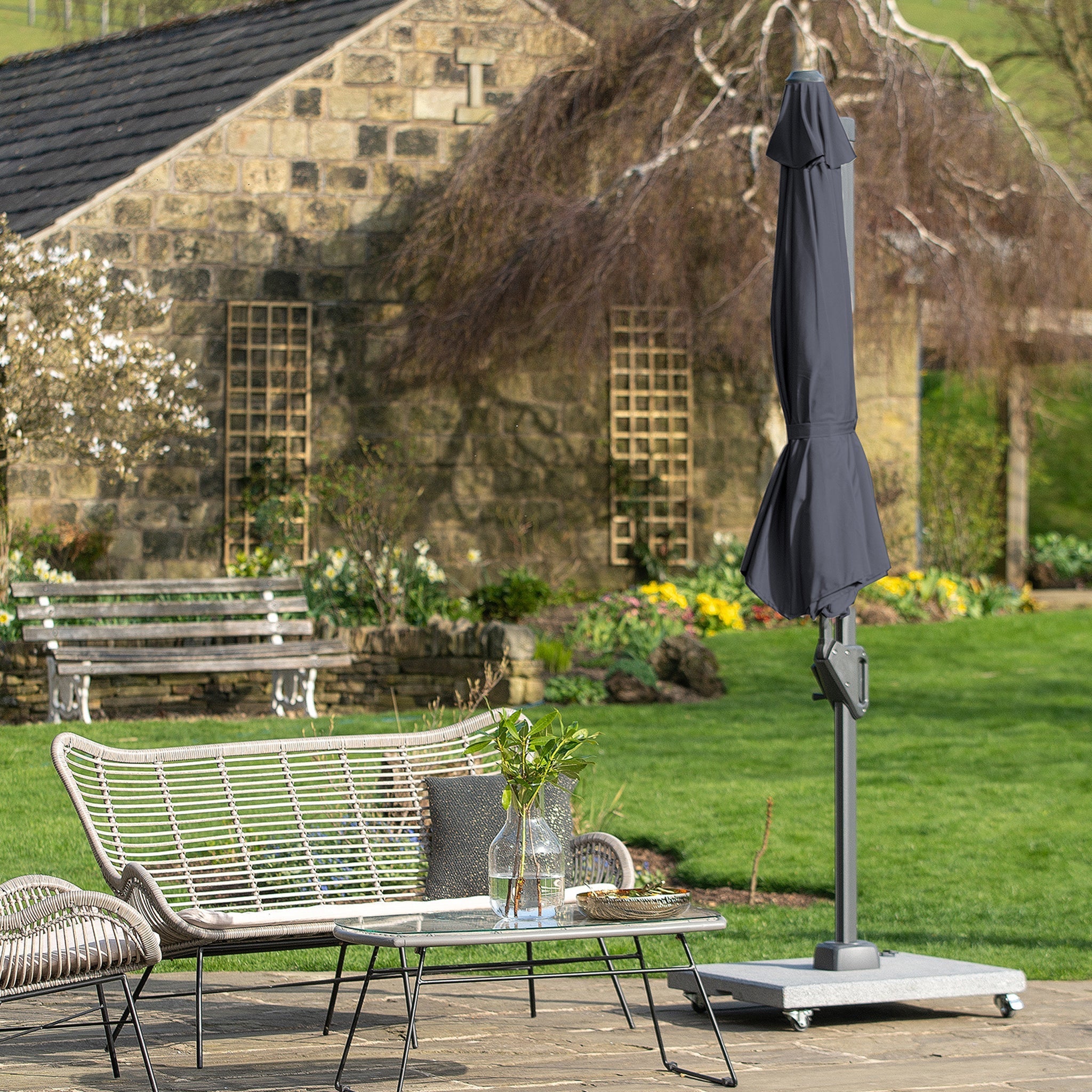 Outdoor garden patio with wicker furniture, a glass-top table, and a Voyager T1 3m Round Parasol in Anthracite; rustic stone building in the background.