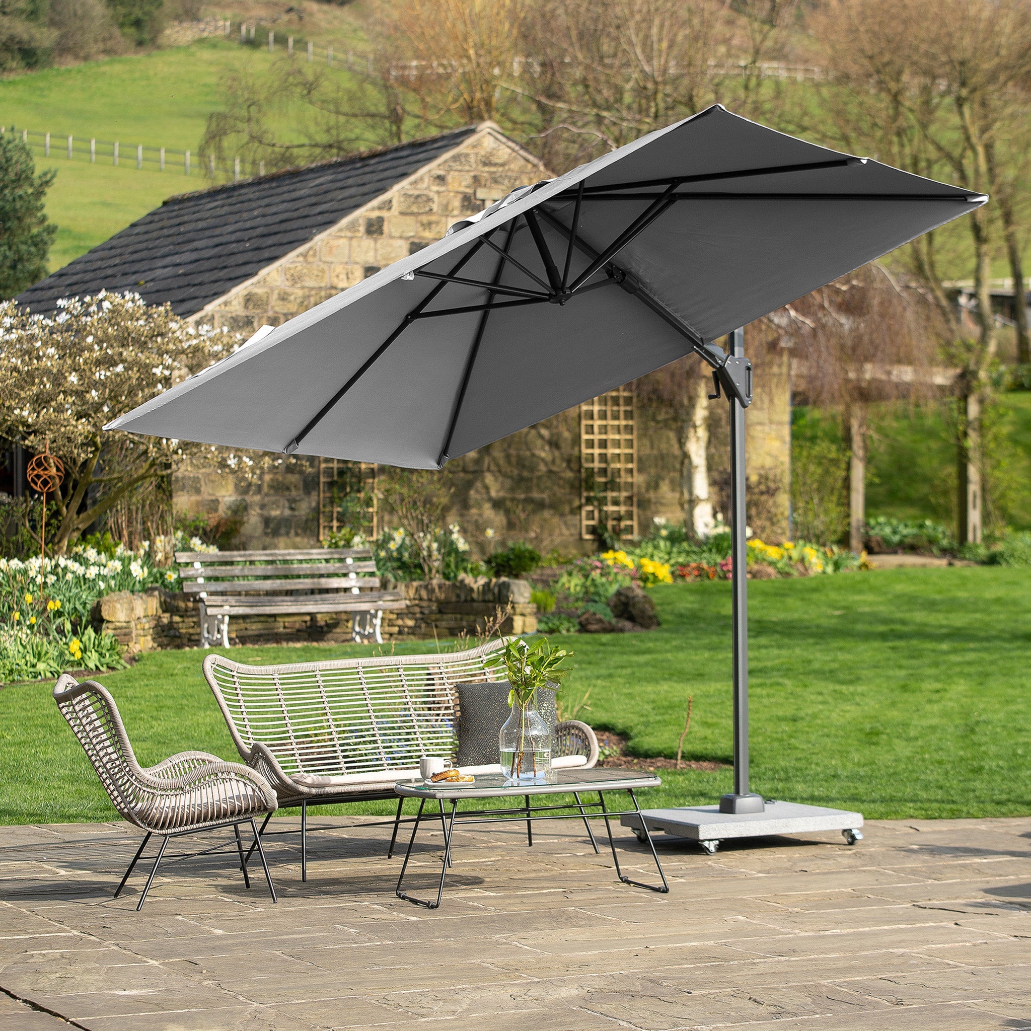 The patio features a Pacific Lifestyle Voyager T2 2.7m Square Parasol in Grey, providing shade to the wicker chairs and table set against a lush garden backdrop.