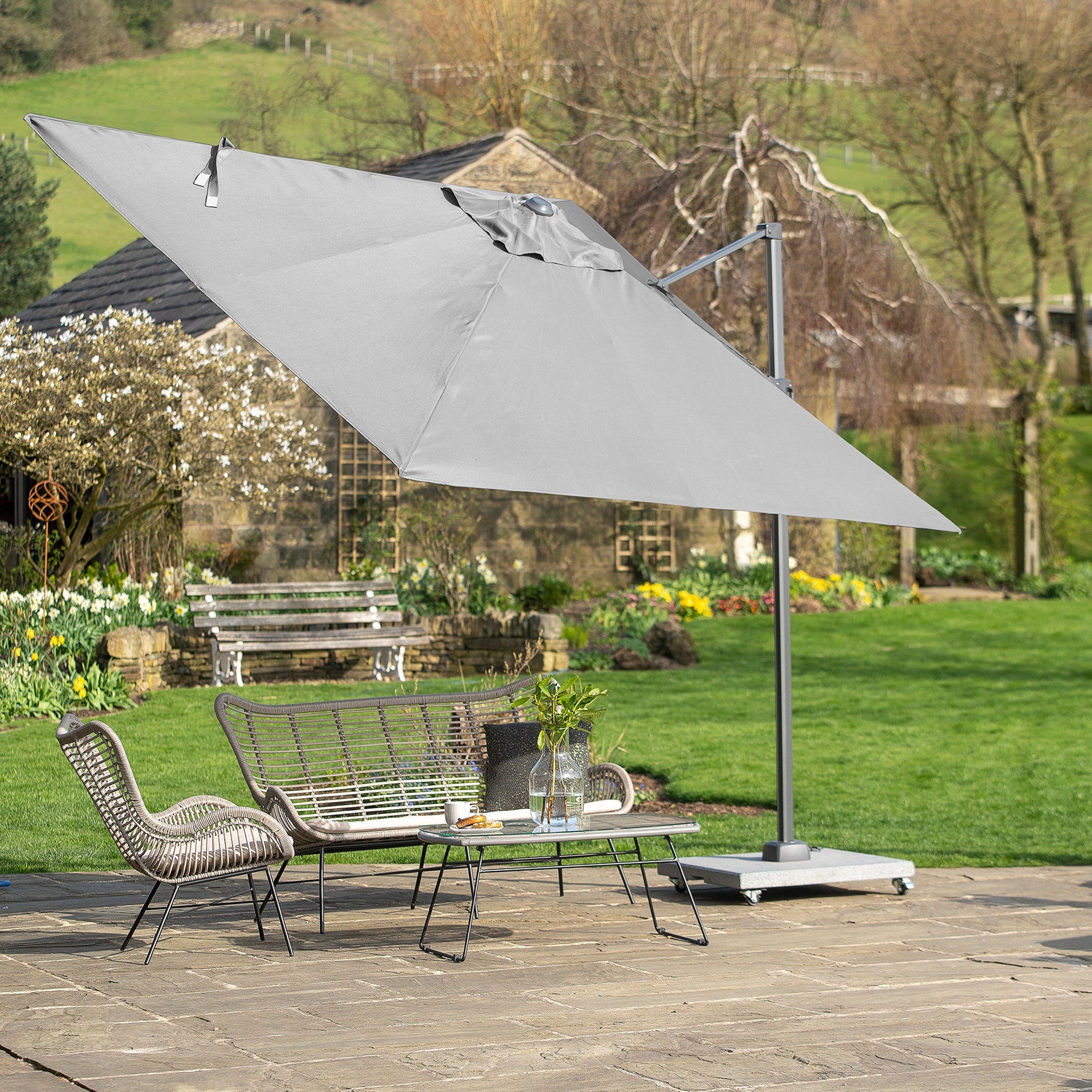Outdoor patio showcasing a Pacific Lifestyle Voyager T2 2.7m square parasol in grey, metal chairs, a table with drinks, and a lush garden backdrop. The parasol provides UPF35+ protection, ideal for comfortable enjoyment on sunny days.