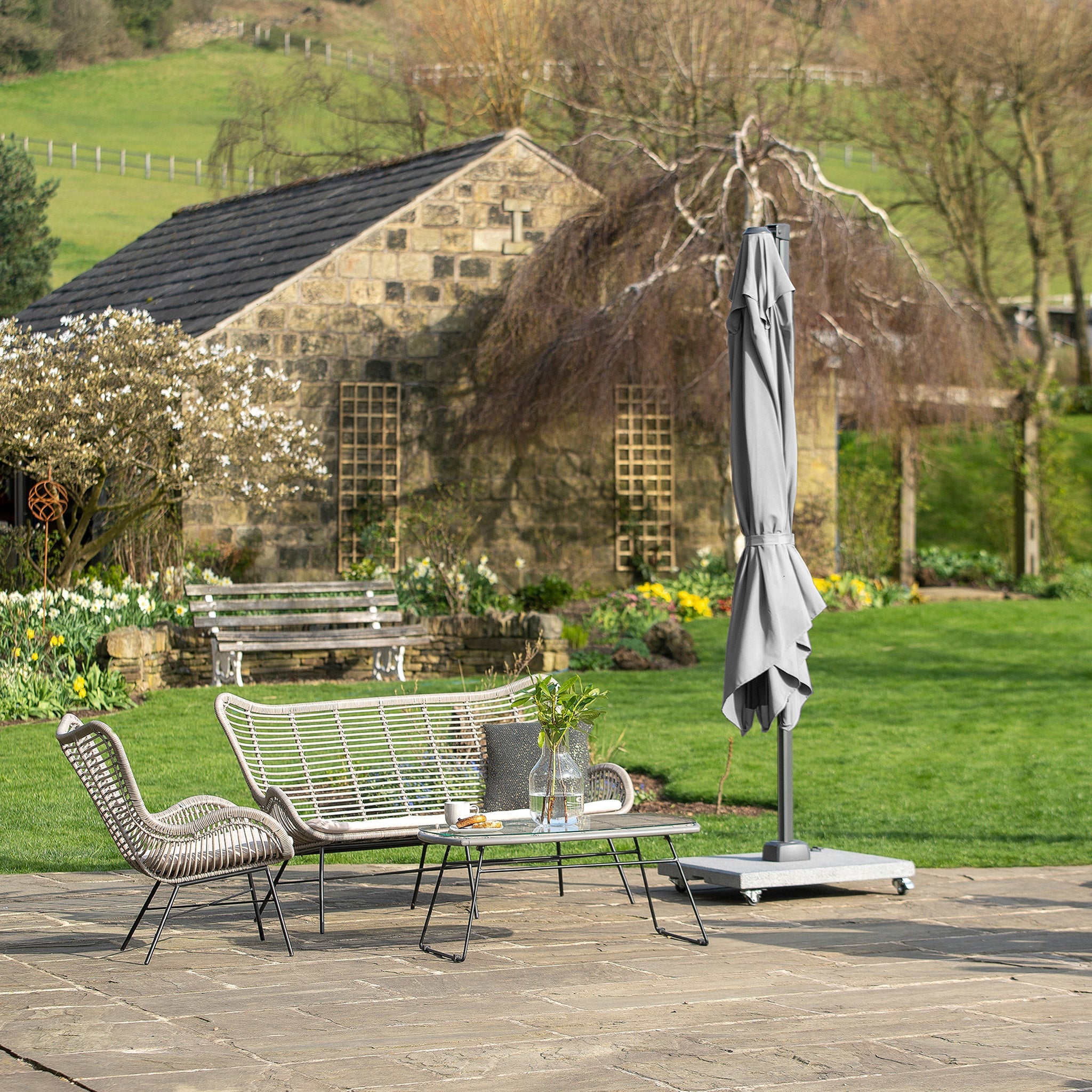Patio featuring wicker furniture, a Pacific Lifestyle Voyager T2 2.7m Square Parasol in Grey, and a stone bench on a paved garden area near a stone house.