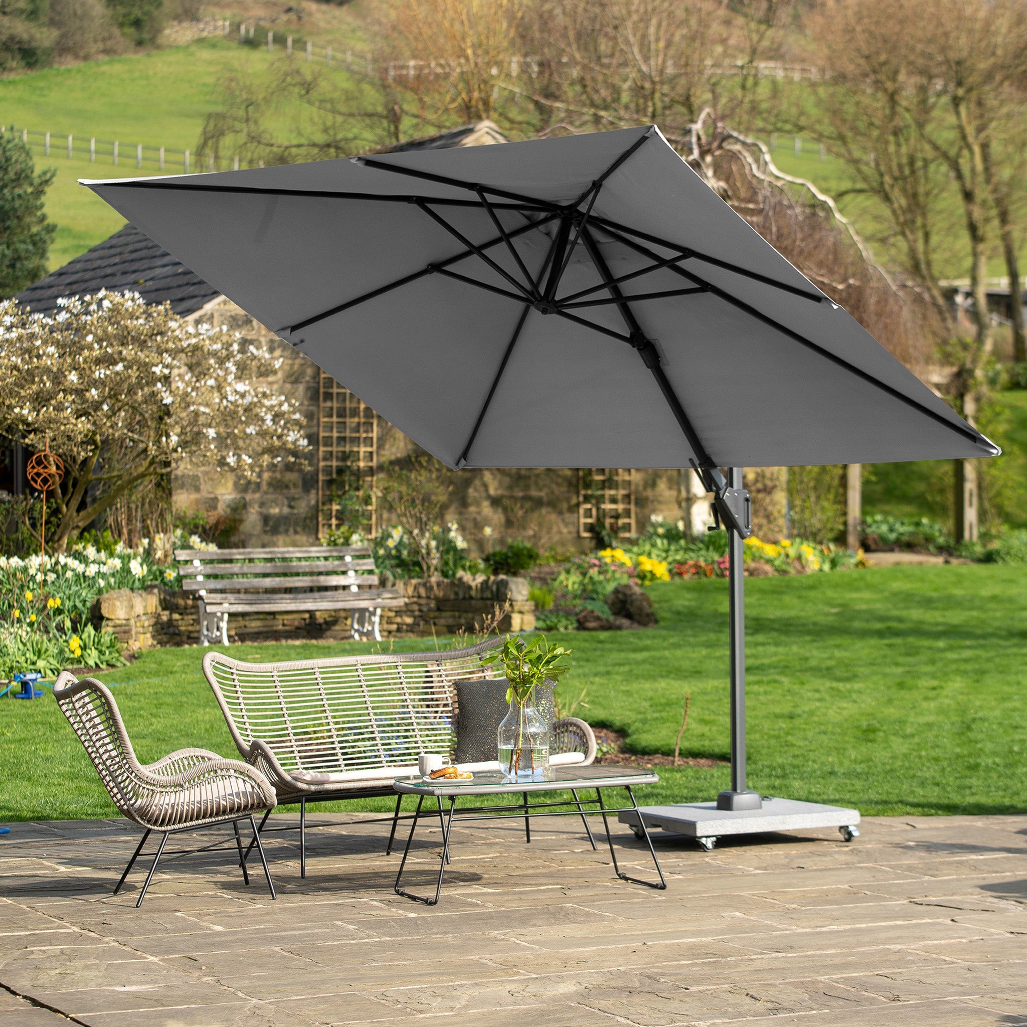 A Pacific Lifestyle - Voyager T2 2.7m Square Parasol in Grey provides shade for wicker chairs and a table on stone tiles, with a garden backdrop.