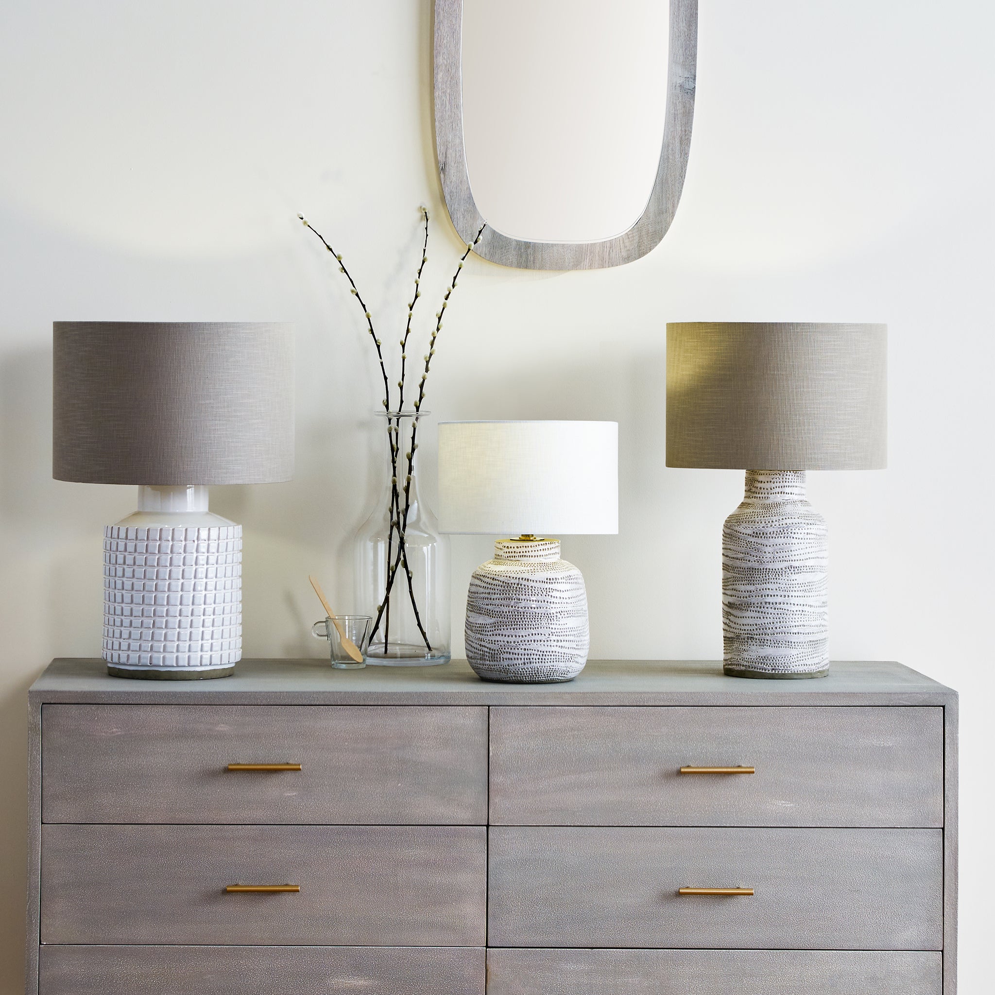 Three Pacific Lifestyle Alina table lamps, featuring white dot designs on stoneware bases, accentuate a wooden dresser adorned with a mirror and a petite pot vase in the background.