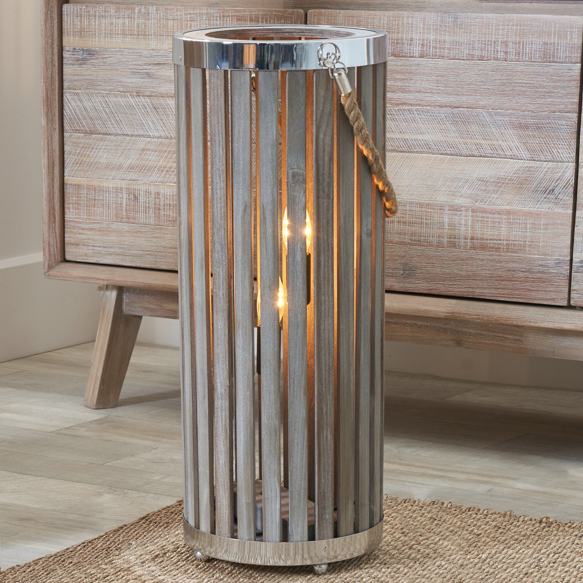 The Pacific Lifestyle - Austell Grey Wash Wood Large Lantern Floor Lamp features a tall cylindrical metal design with vertical slats and a rope handle, casting a warm glow on its grey wash wood base and exuding coastal charm.
