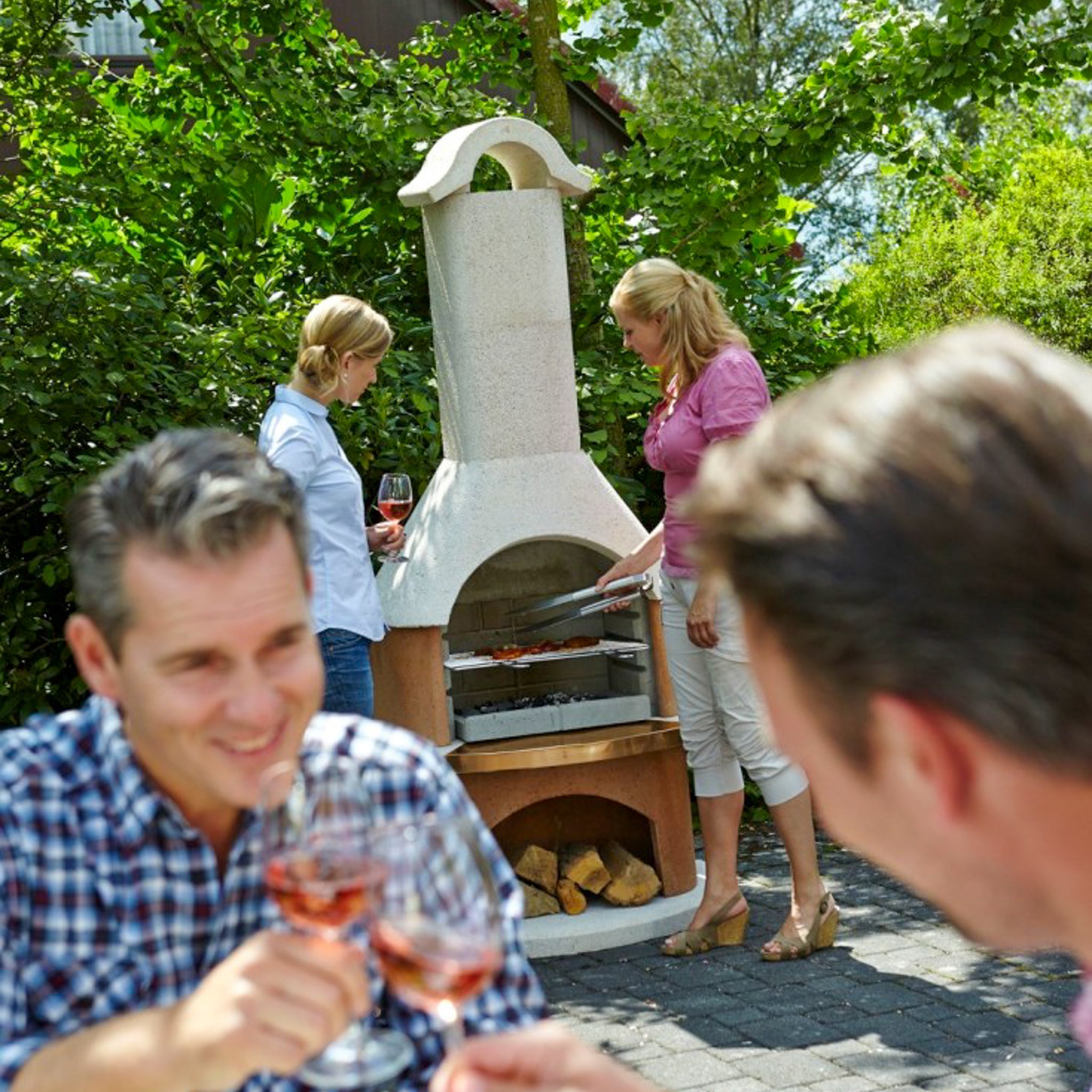 People enjoying wine near an outdoor fireplace as two women prepare food on the grill, using the elegant Buschbeck Bologna Masonry Barbecue with its sophisticated white quartz structure.