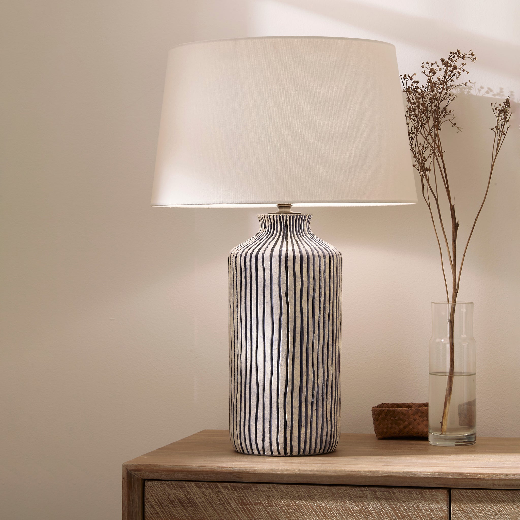 The Pacific Lifestyle - Bude Blue and White Stripe Stoneware Table Lamp rests on a wooden table, harmonizing with the coastal decor beside dried flowers in a glass vase.