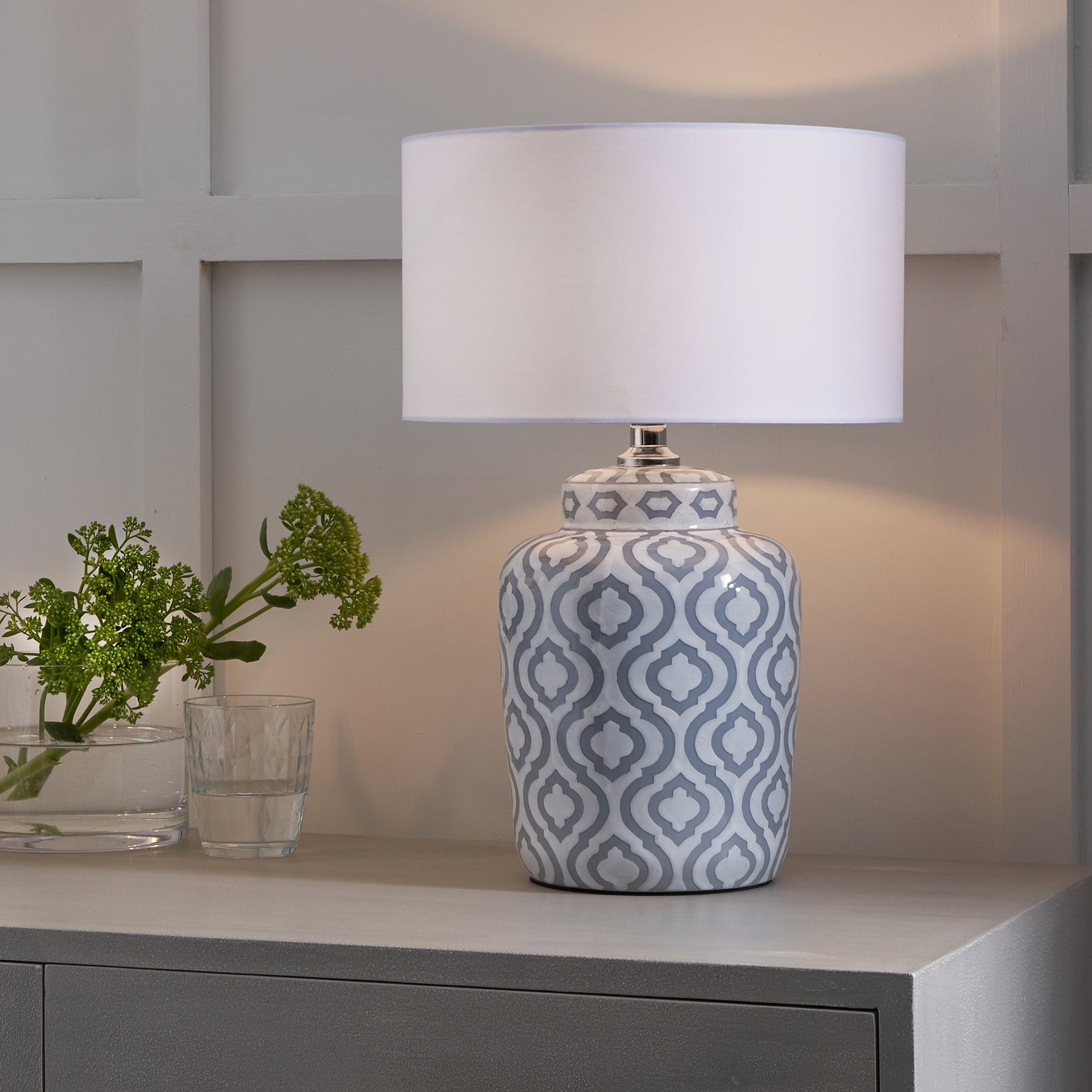 The Pacific Lifestyle Celia Grey and White Pattern Ceramic Table Lamp, with its geometric design and white shade, sits elegantly on a cabinet next to a vase of green flowers, enhancing the setting's charm.