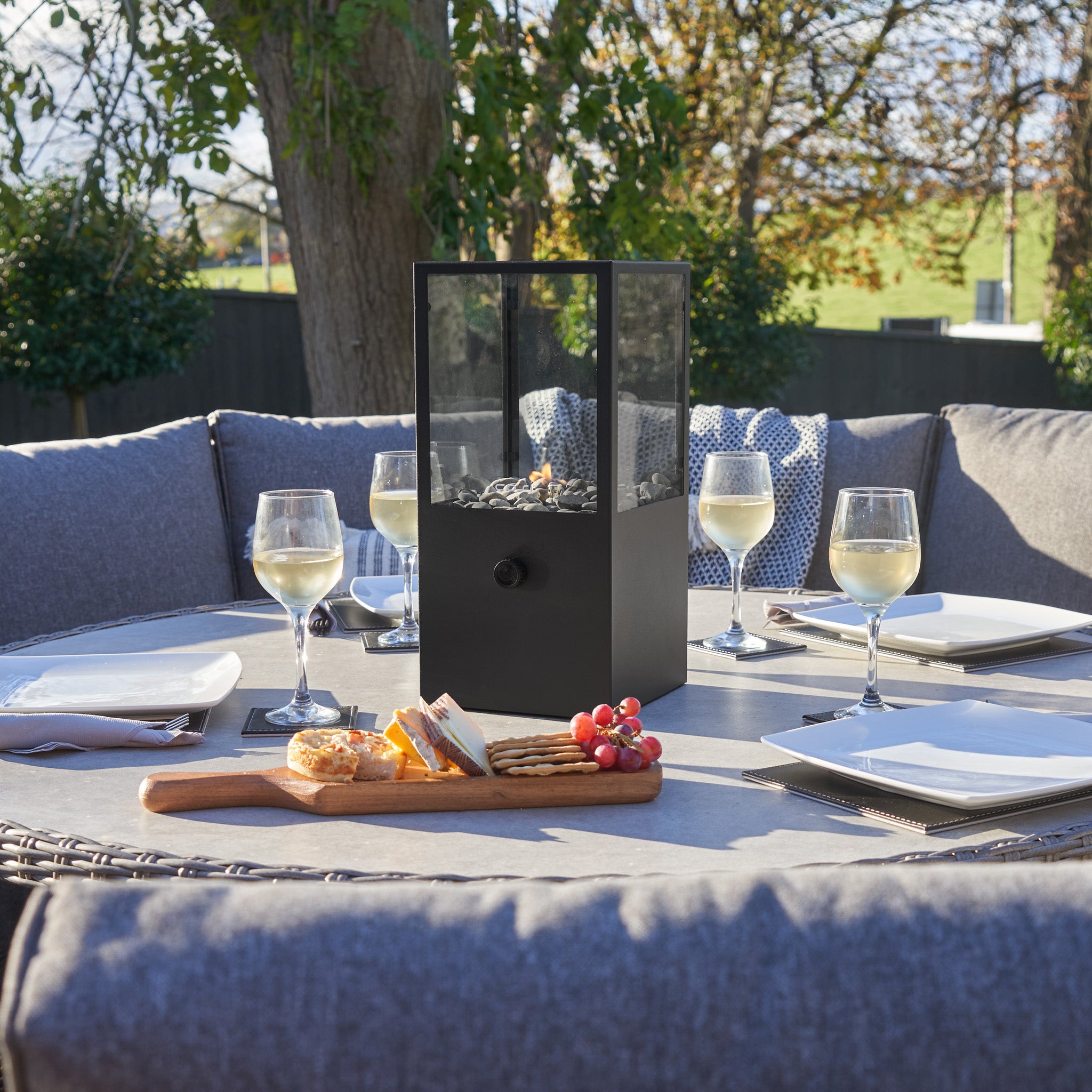 An outdoor dining table features wine glasses, plates, and a black centerpiece. Complemented by the Cosi Cosidome High Black Fire Lantern, cheese and grapes rest invitingly on a cutting board.