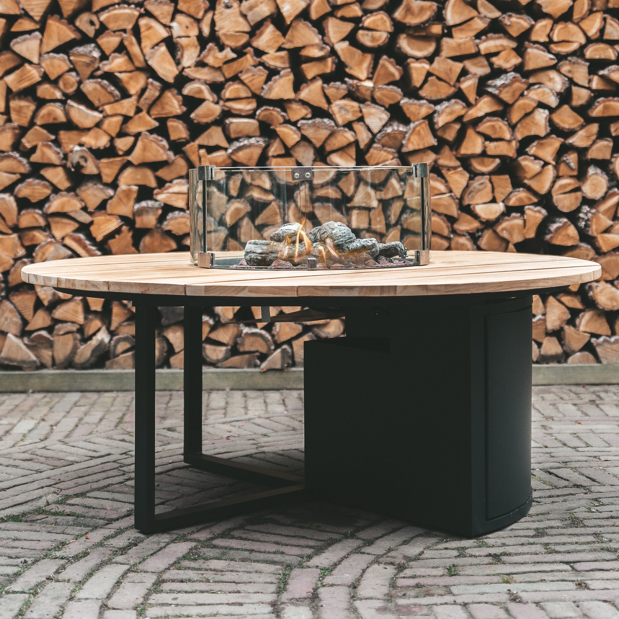 The Cosi - Cosiloft 120 Black and Teak Round Fire Pit is a round outdoor table with a teak top and glass-enclosed fire pit, stylishly placed on a brick patio against stacked wood.