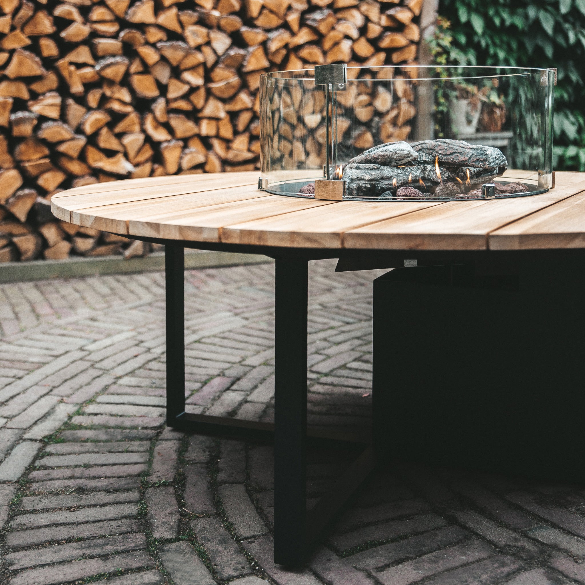 The Cosi - Cosiloft 120 Black and Teak Round Fire Pit features a teak wood top with a glass-enclosed center, perfect for cozy evenings. It pairs beautifully with the brick surface and stacked wood background.