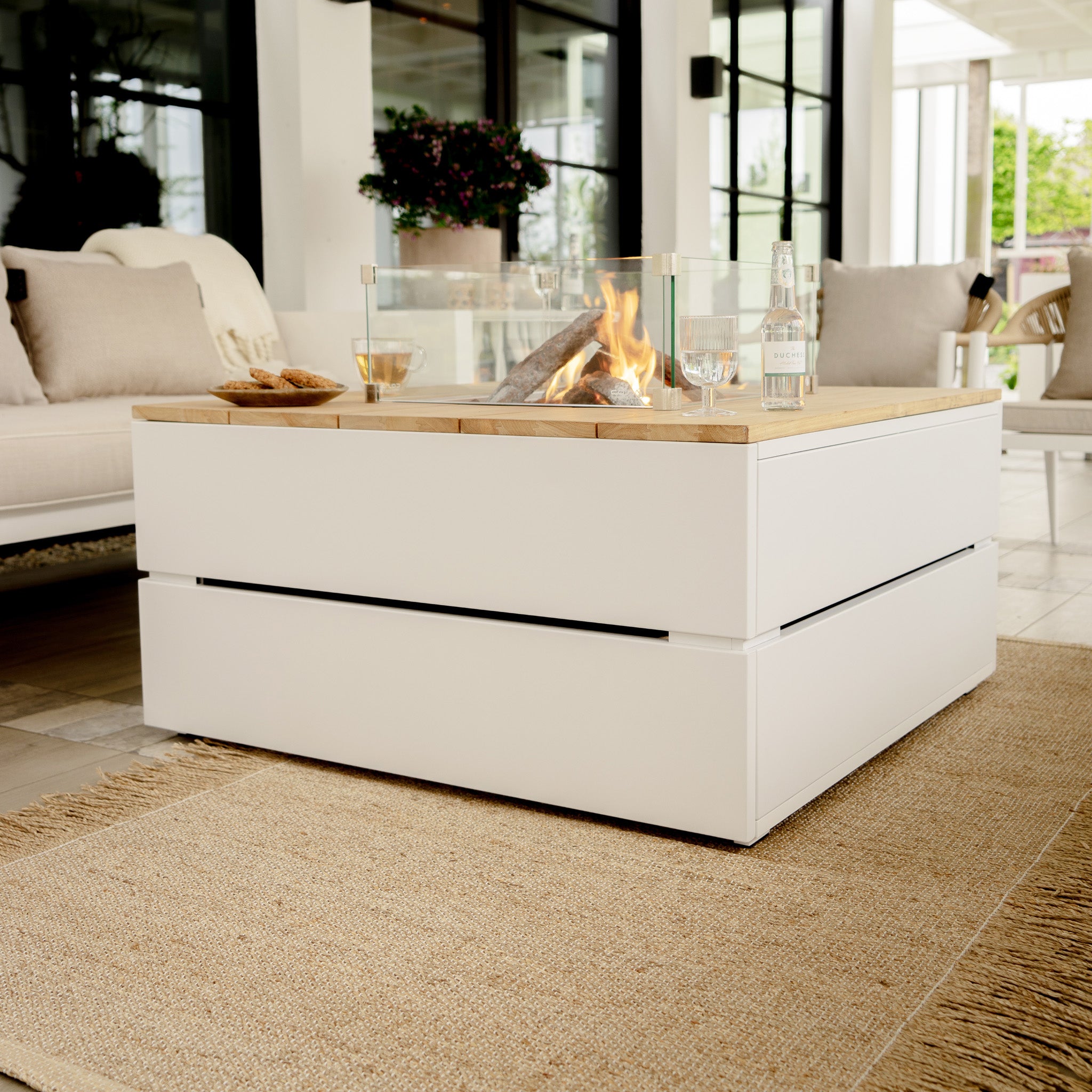 A modern patio showcases the Cosi - Cosipure 100 White and Teak Square Fire Pit as its centerpiece, surrounded by beige furniture and a woven rug, epitomizing sophisticated garden design.