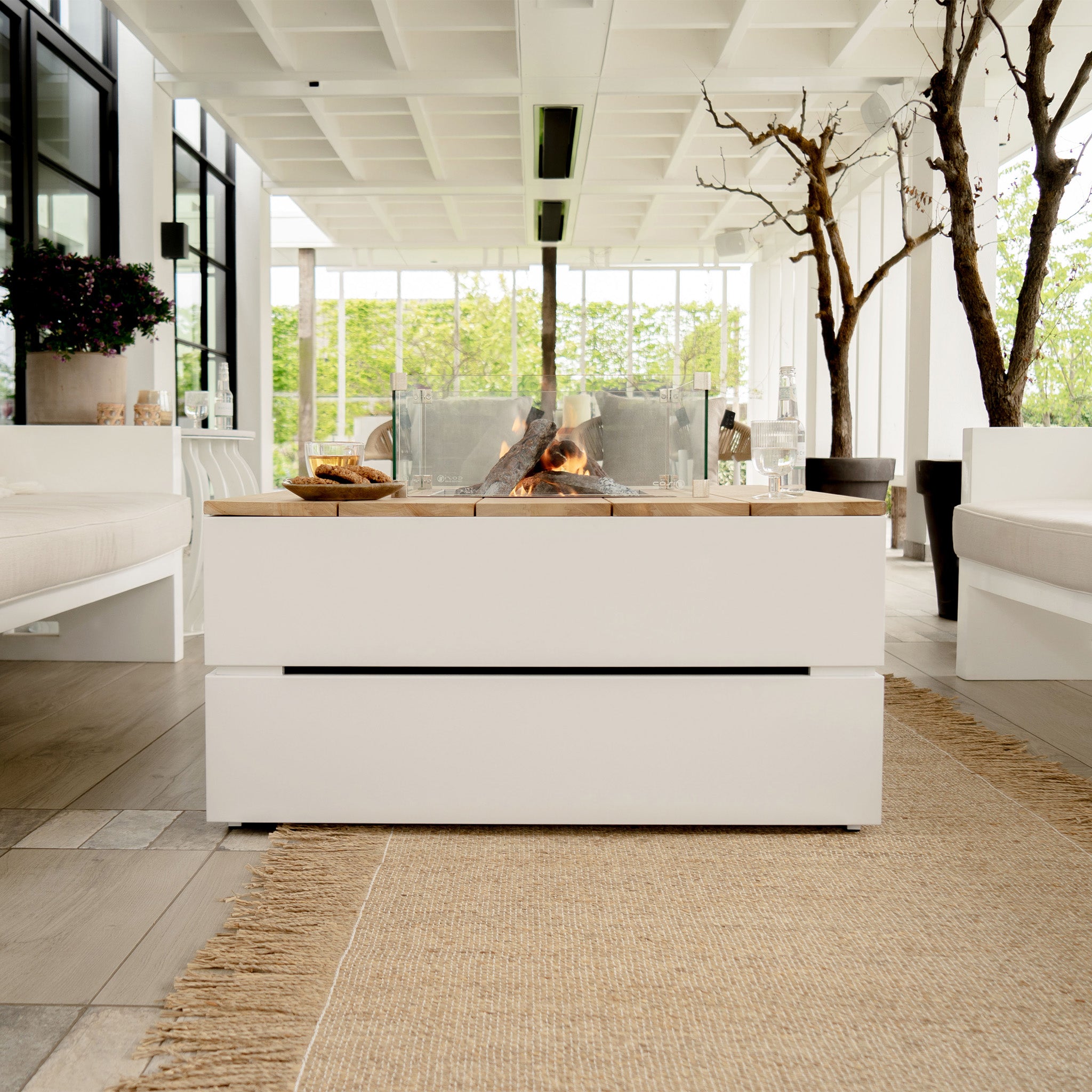 A modern outdoor patio featuring the Cosi - Cosipure 100 White and Teak Square Fire Pit, complemented by white seating and potted plants beneath a geometric ceiling, seamlessly blending style and garden design.