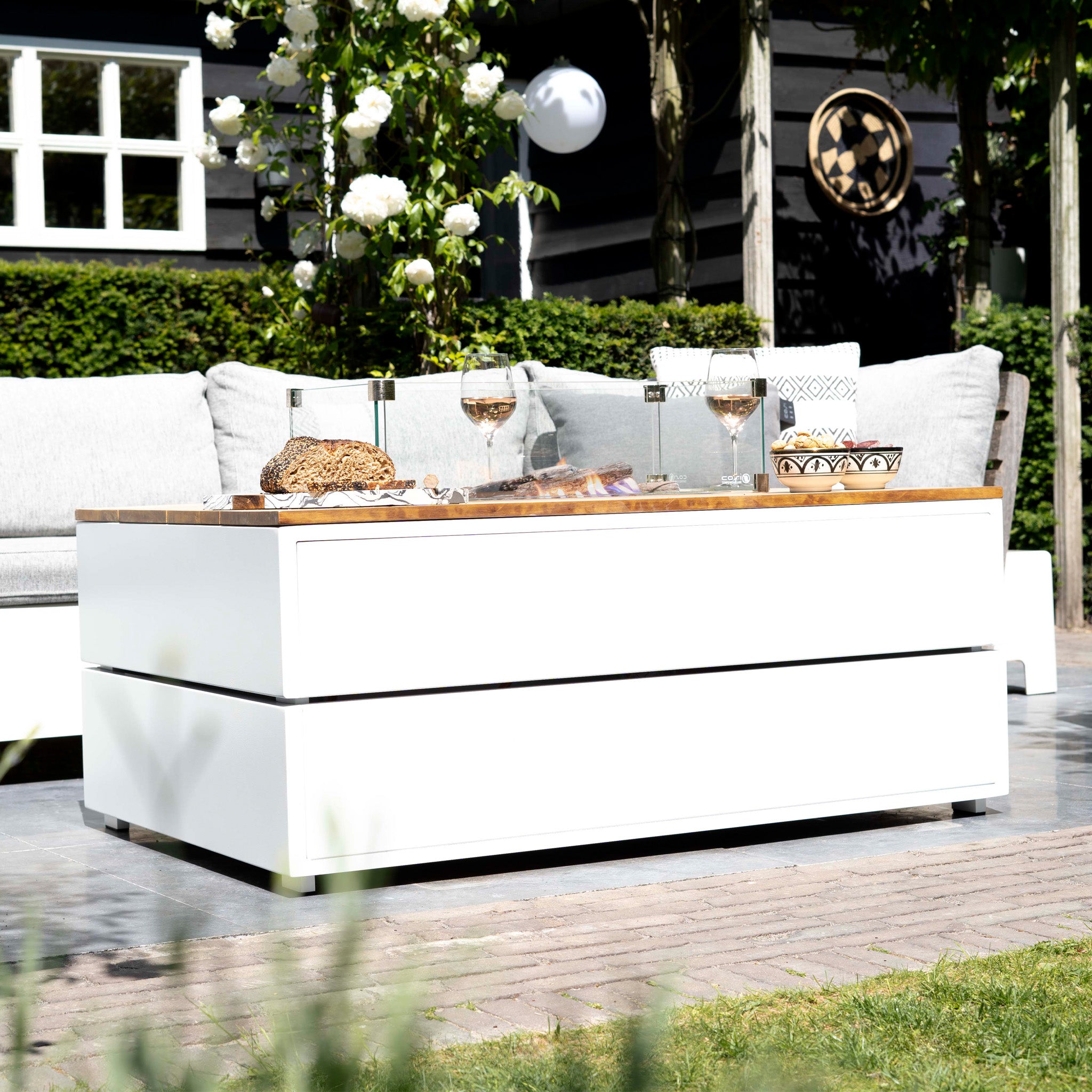 An outdoor seating area includes wine glasses and snacks on a white table, enhanced by the Cosi - Cosipure 120 White and Teak Rectangular Fire Pit. The backdrop of a black house and lush greenery adds an inviting ambiance for any gathering.