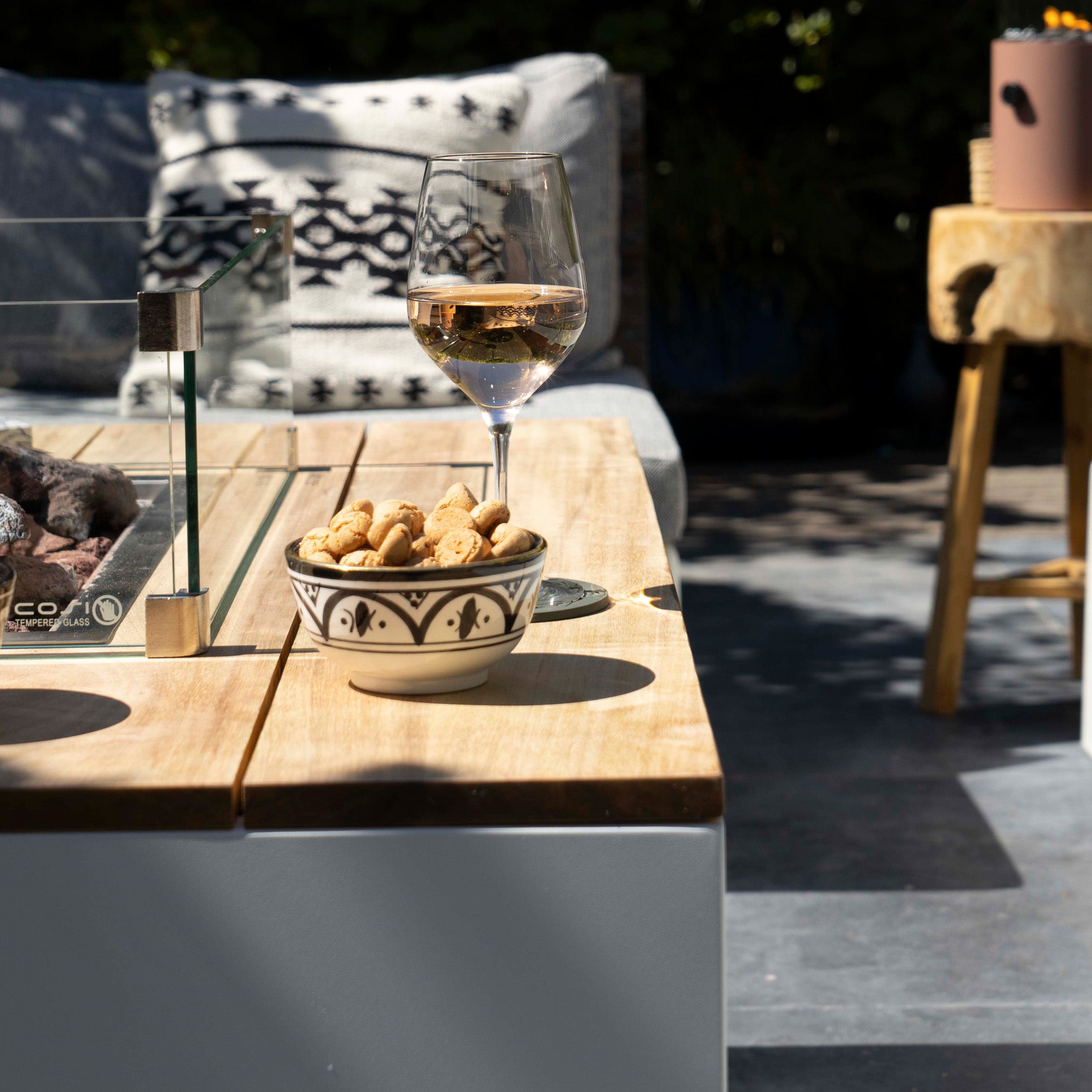 A glass of rosé wine and a bowl of peanuts sit on a wooden table next to the warm glow of the Cosi - Cosipure 120 White and Teak Rectangular Fire Pit, creating a cozy setting.
