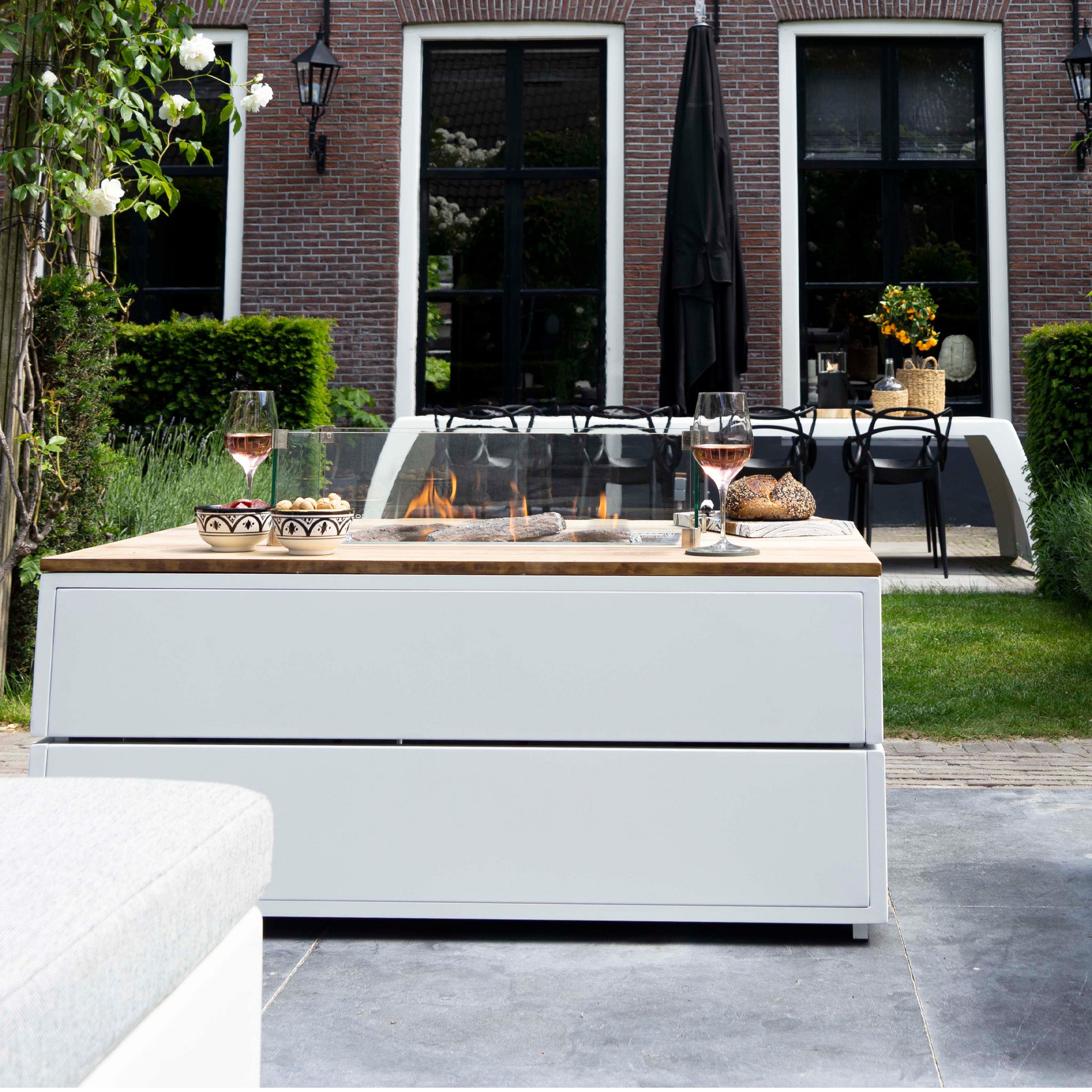 Outdoor patio featuring the Cosi - Cosipure 120 White and Teak Rectangular Fire Pit, wine glasses, and dining setup in front of a brick house.