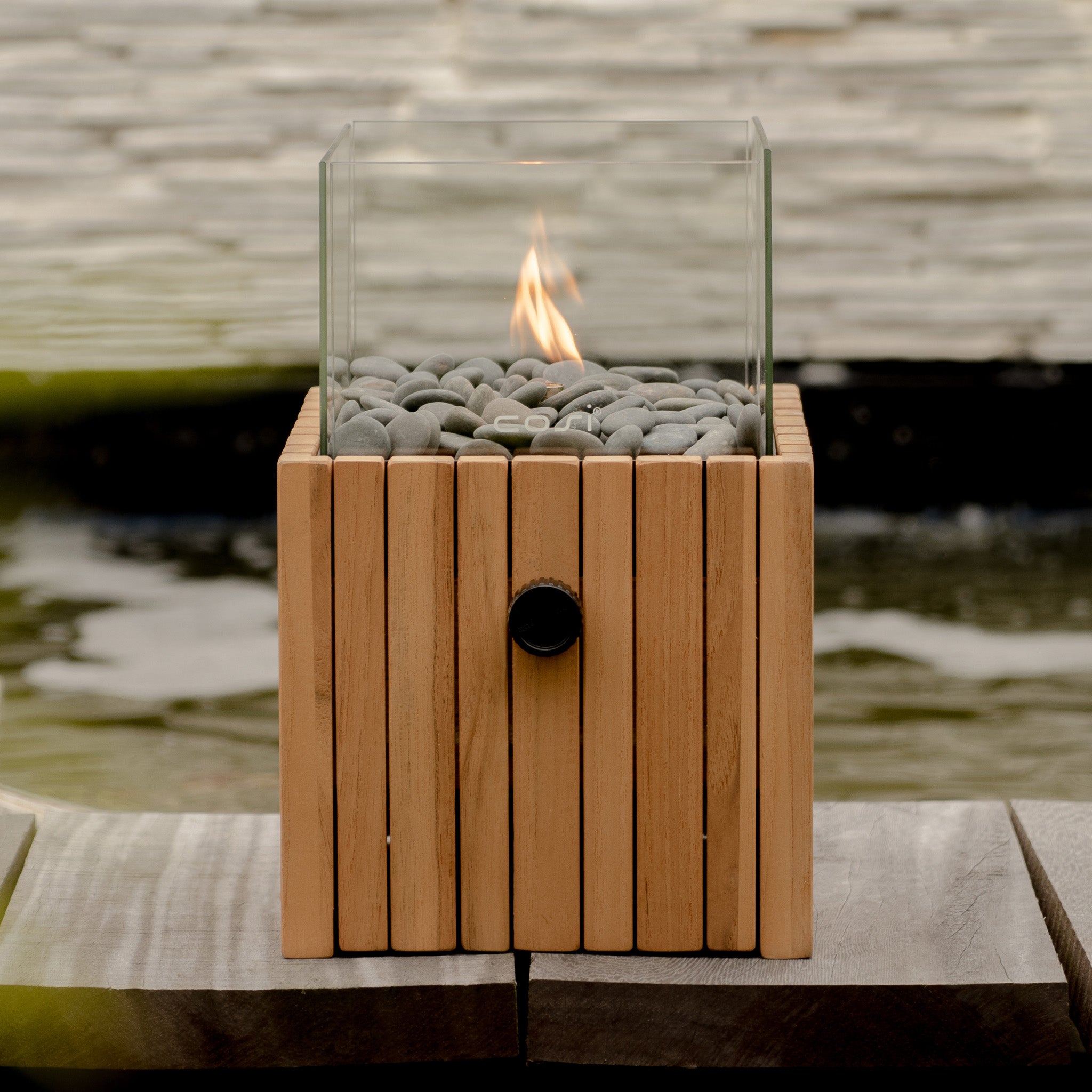 The Cosi - Cosiscoop Timber Square Fire Lantern, with glass sides and gray stones, sits elegantly on wood by the water feature. It creates a captivating atmosphere and combines style with functionality for your outdoor space, effortlessly powered by a gas cartridge.