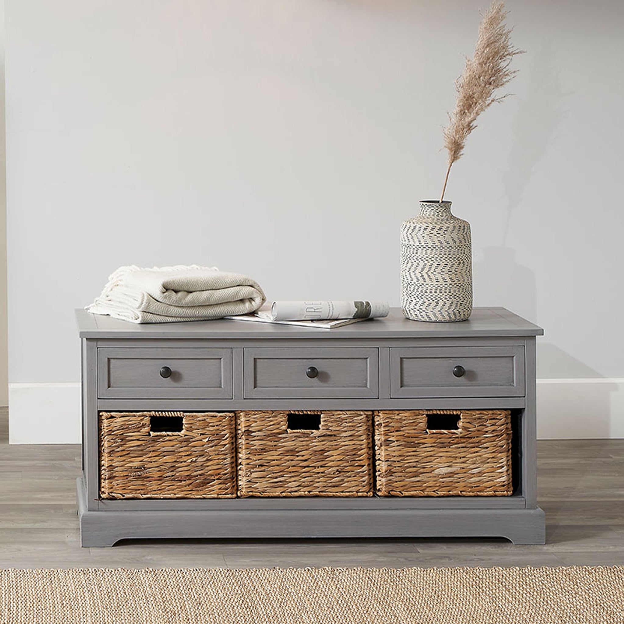Part of the Devonshire collection, the Pacific Lifestyle - Devonshire Grey Wood 3 Drawer 3 Basket Unit offers a stylish storage solution with its gray wash finish, topped with a vase, rolled gray blanket, and magazine.