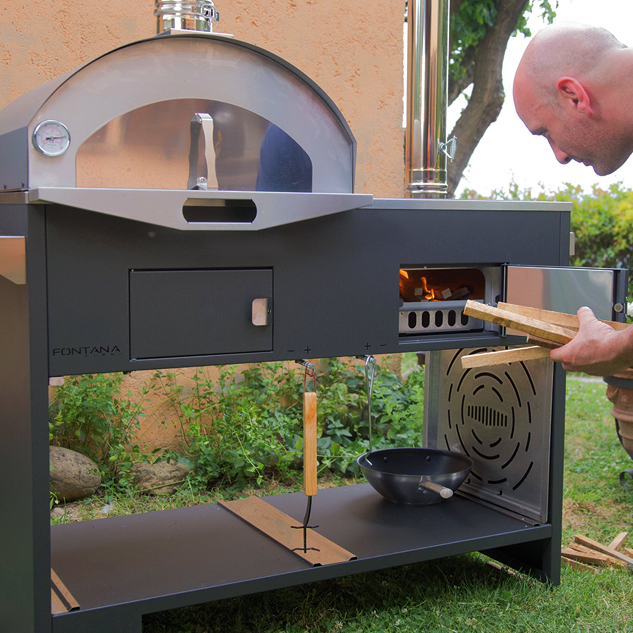 Person placing wood into a modern Fontana Pizza & Cucina Oven Including Trolley, with a fire burning inside, set in a garden.