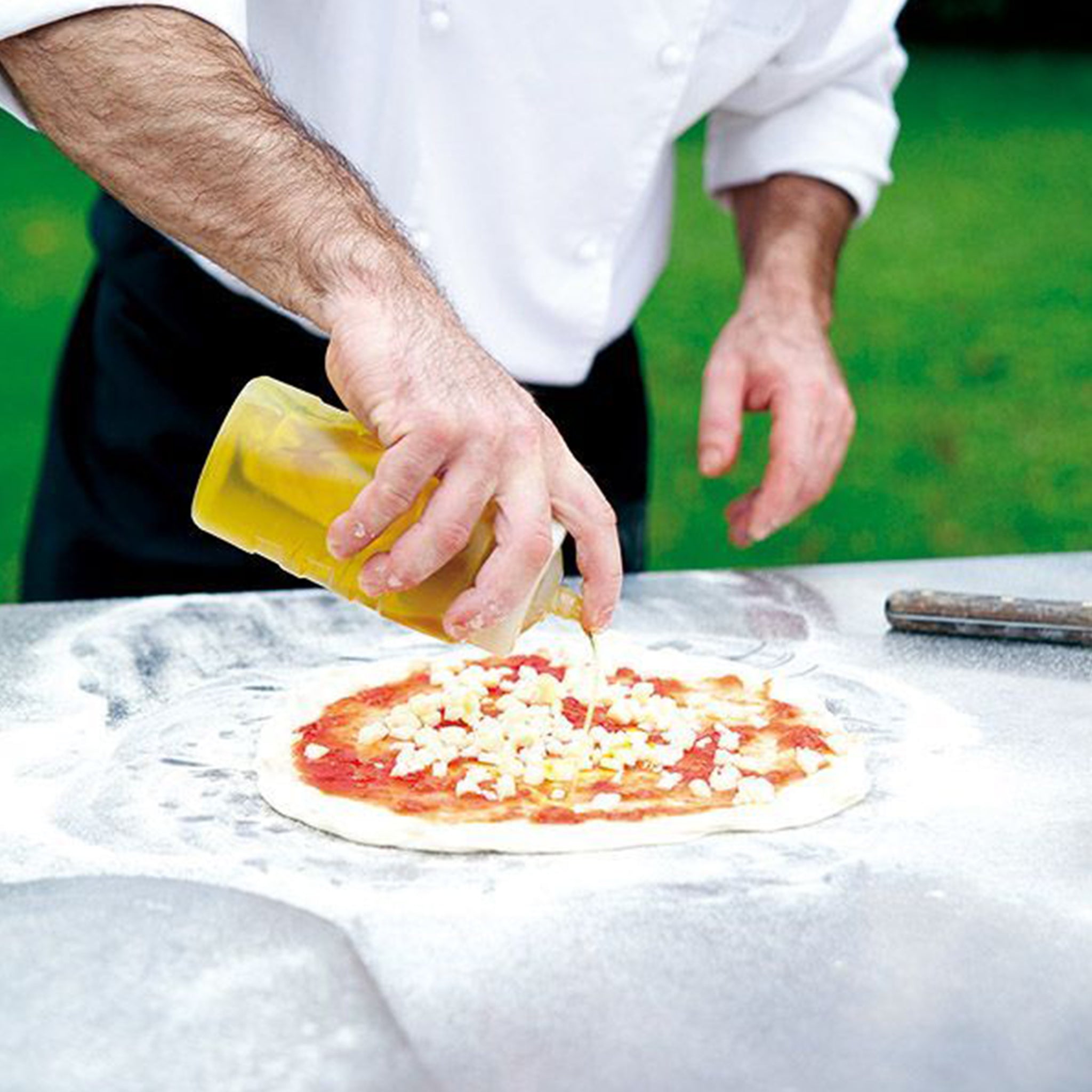 A person in a white chef jacket, standing next to a Fontana Capri Wood Pizza Oven, drizzles olive oil on a pizza dough topped with cheese and tomato sauce.