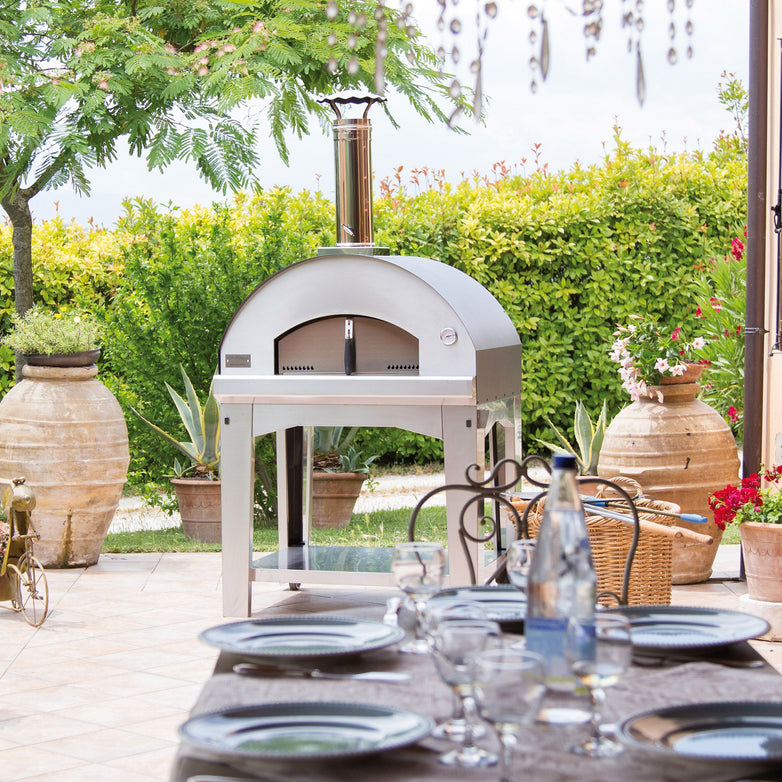 Outdoor patio featuring the Fontana Mangiafuoco Stainless Wood Pizza Oven Including Trolley, a tastefully arranged dining table set with plates and glasses, all embraced by lush greenery.