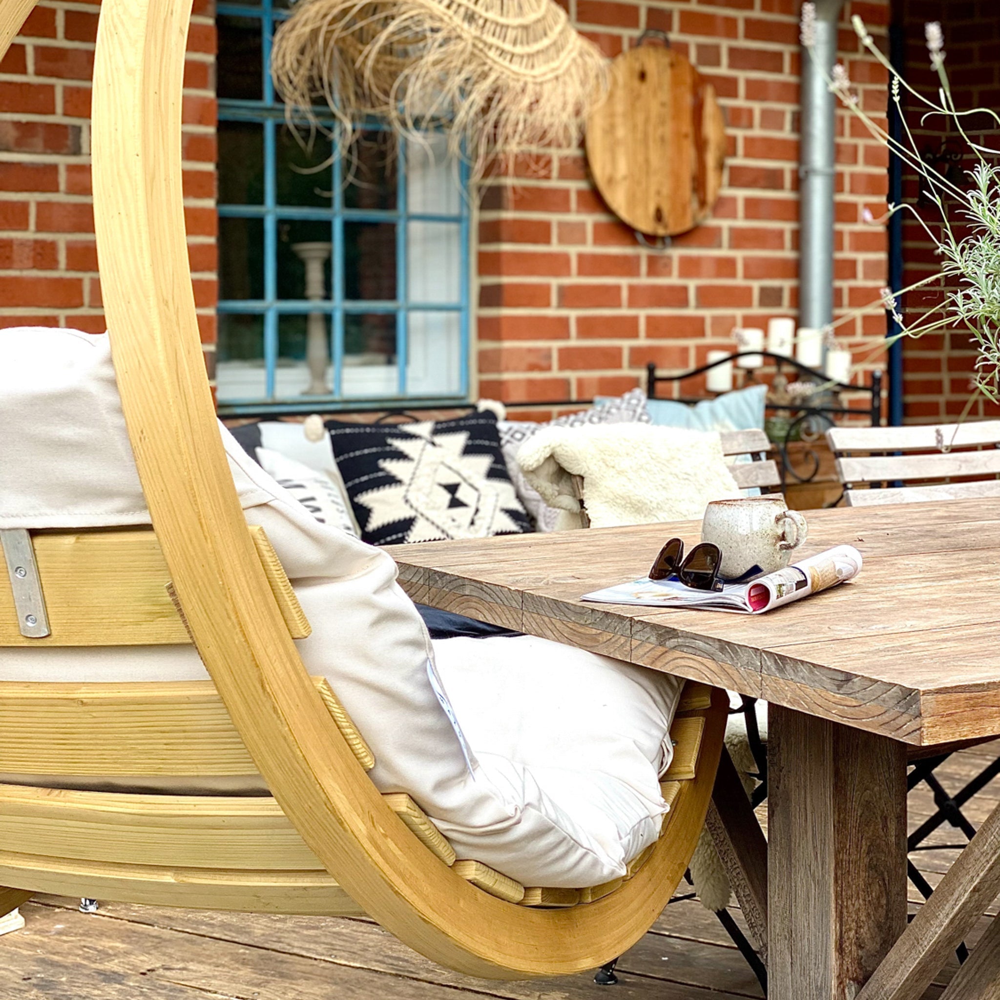 A cozy patio featuring the Swing Lounger in Creme, a wooden table adorned with pillows, a coffee mug, and a rolled magazine on a sunny day.