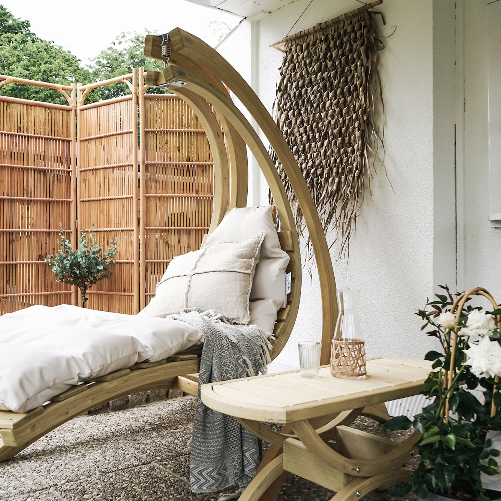 Swing Lounger in Creme with water-repellent cushions, accompanied by a small table, wicker wall decor, and a bamboo privacy screen.