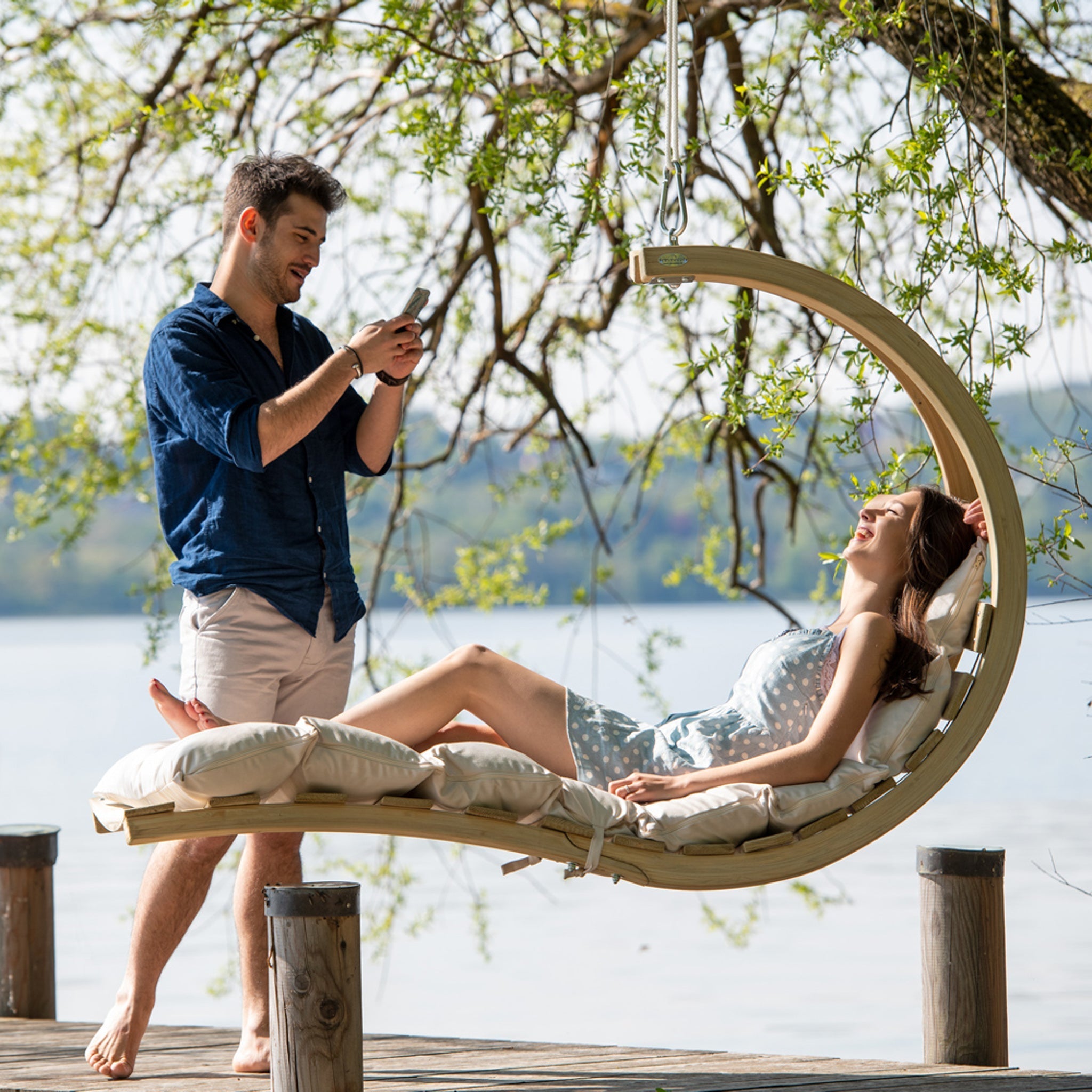 A woman relaxes on the Swing Lounger in Creme by the water as a man stands nearby, capturing the moment with his phone.