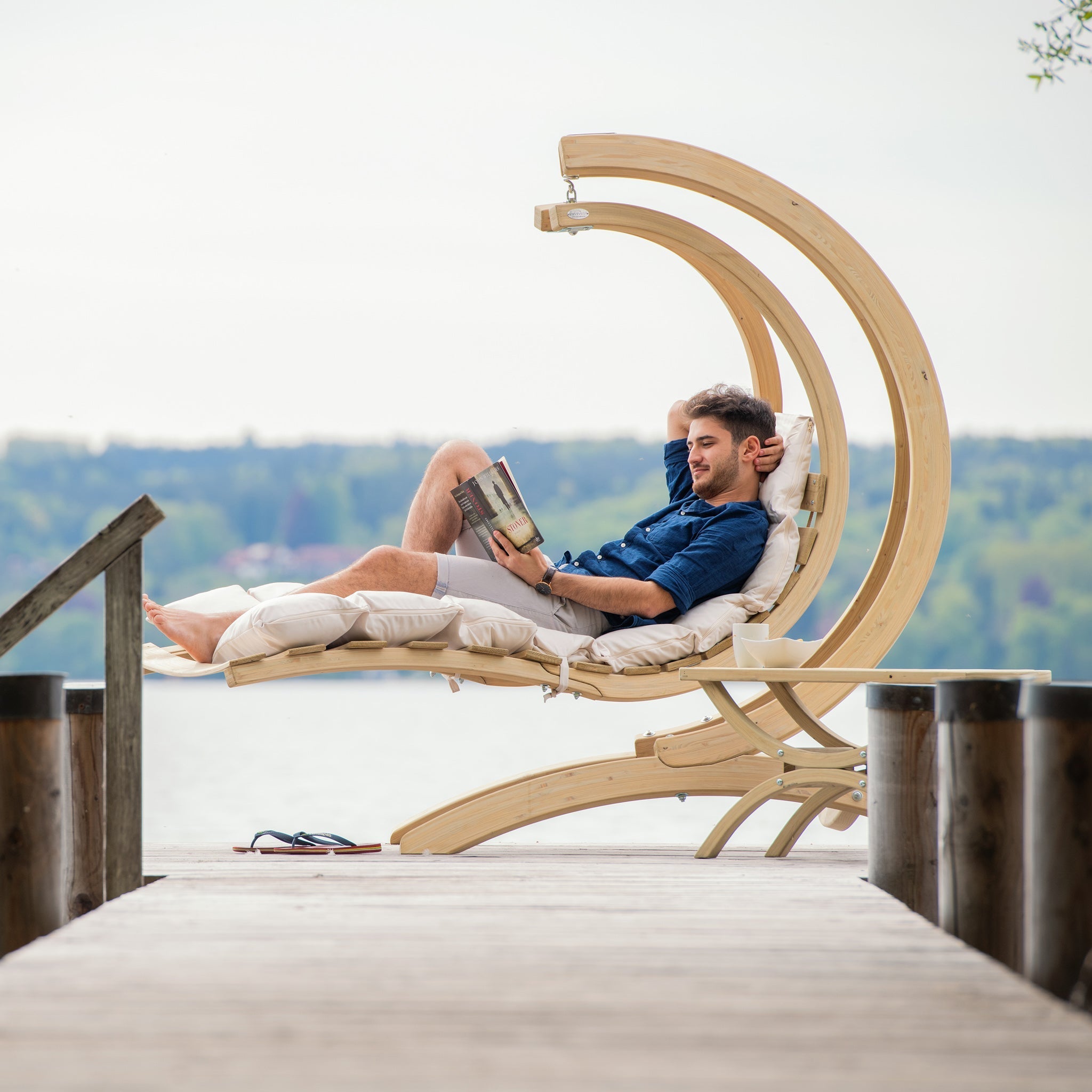 A man relaxes in a Swing Lounger in Creme, enjoying his book by the lakeside pier.