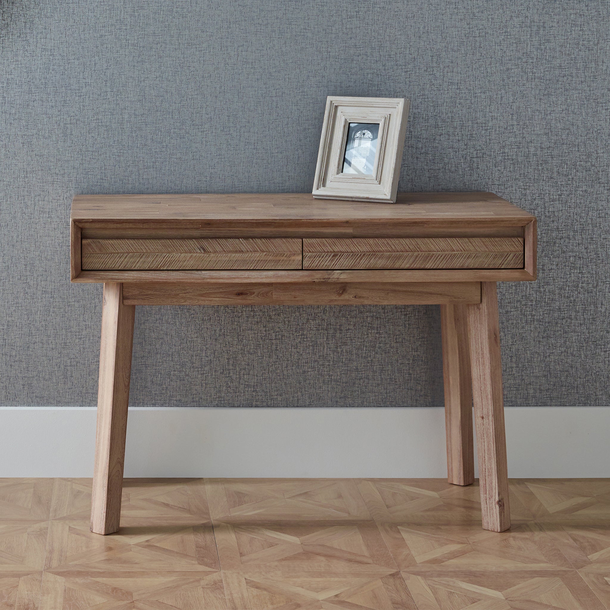 Part of the Kalmar Collection, the Pacific Lifestyle - Kalmar Sand Wash Acacia Wood 2 Drawer Desk embodies Scandi style with sleek lines and two drawers. It features a framed photo on top, placed against a textured gray wall for a minimalist yet warm look.