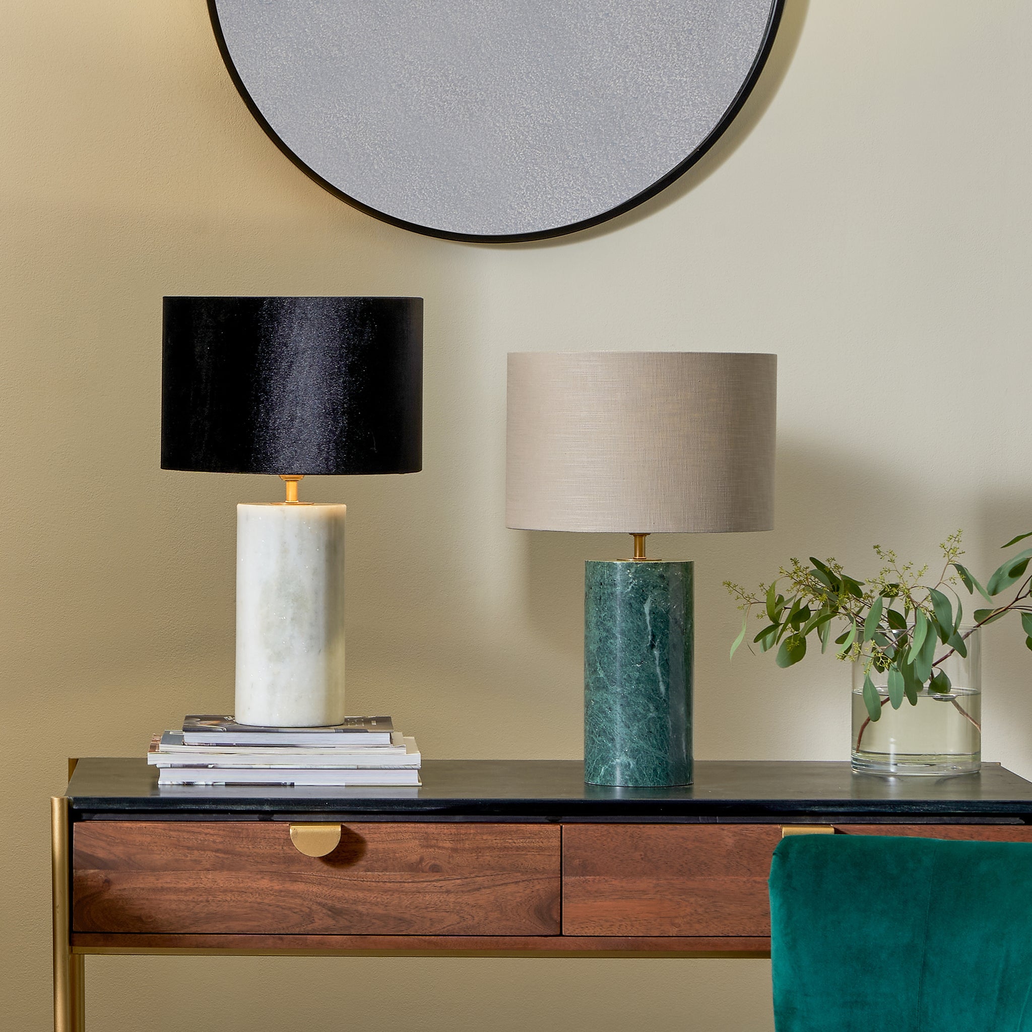 Two lamps with sleek finishes sit on a wooden table, accompanied by a round mirror above and a glass vase with greenery beside them. A Pacific Lifestyle Kiorini White Marble Table Lamp adds elegance to the scene.