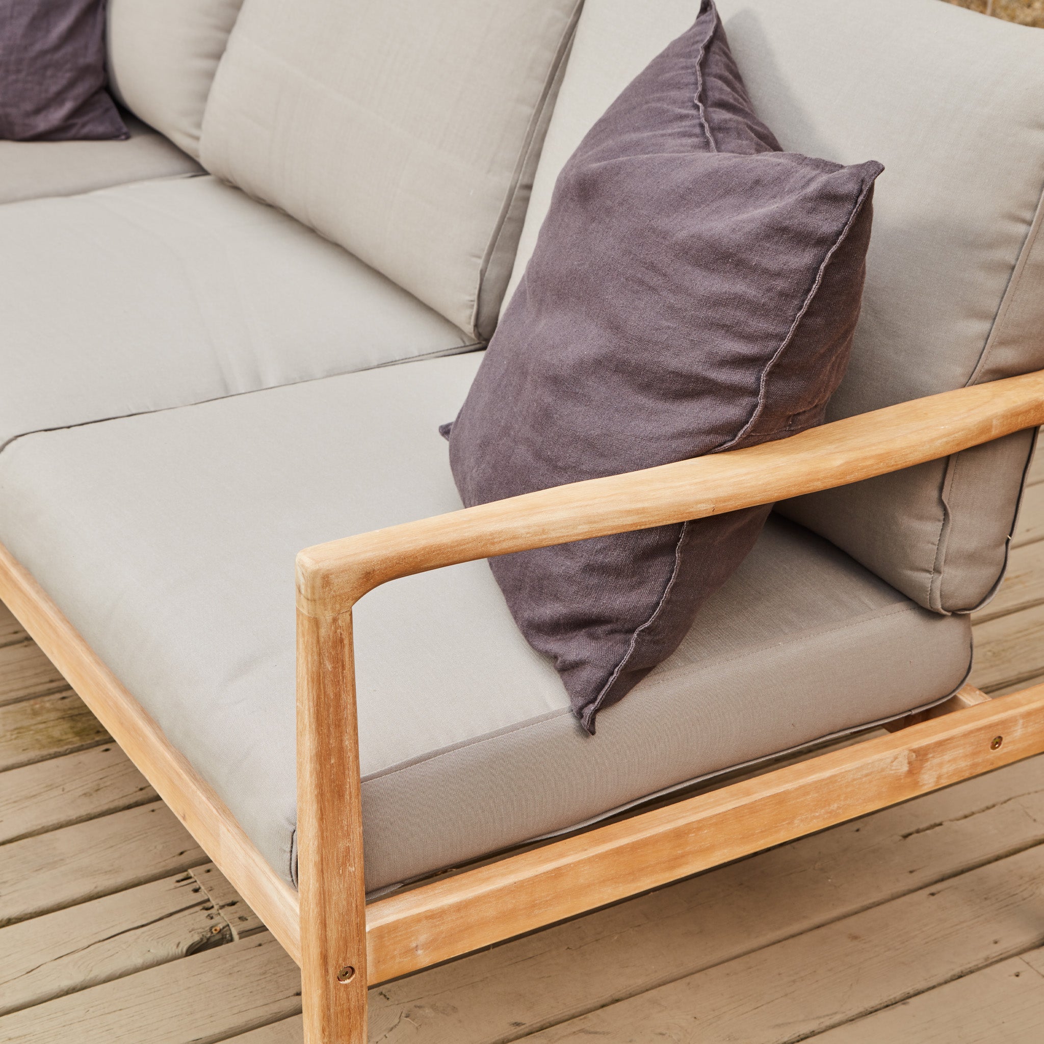 The Pacific Lifestyle Malta Lounge Corner Set in Natural Taupe includes a wooden outdoor sofa with an acacia wood frame, beige cushions, and a vibrant purple pillow, offering a chic oasis on the deck.