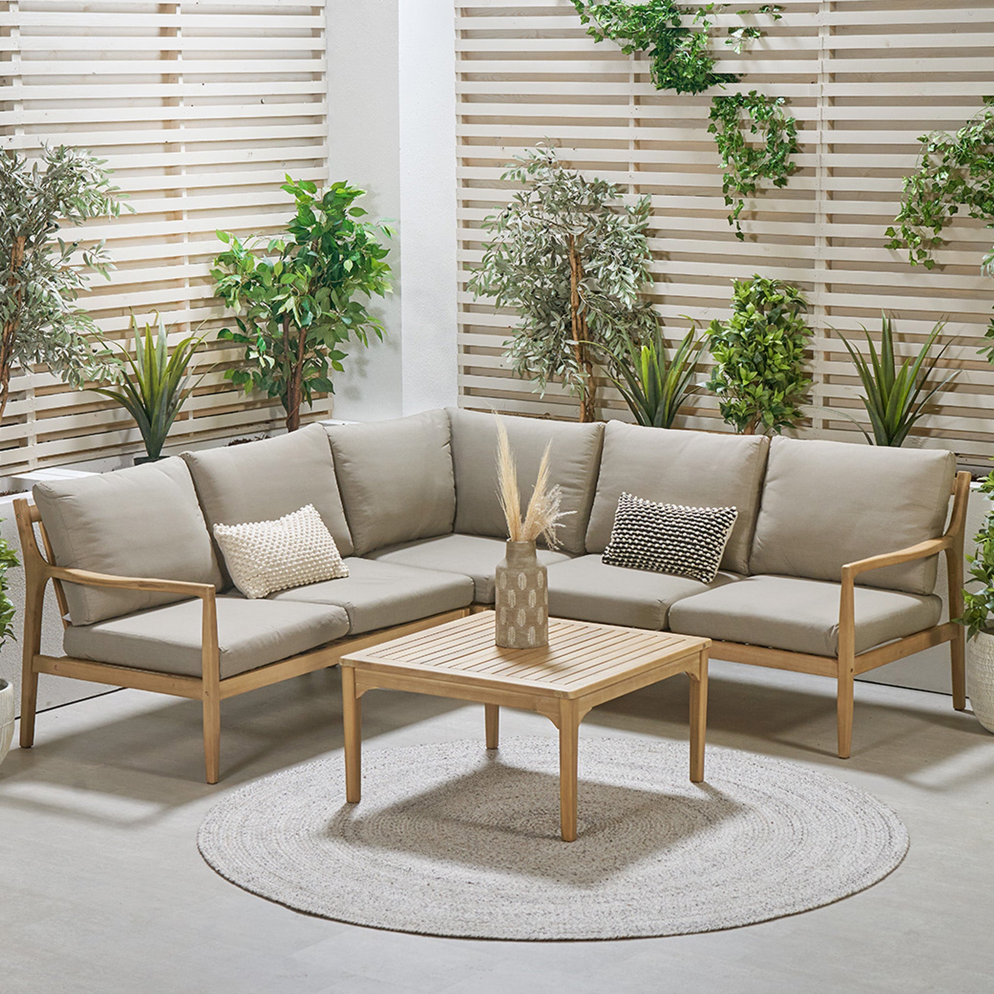 Outdoor patio showcasing the Malta Lounge Corner Set in Natural Taupe by Pacific Lifestyle, paired with a wooden table framed in acacia wood and vibrant green plants against spotless white slatted walls.