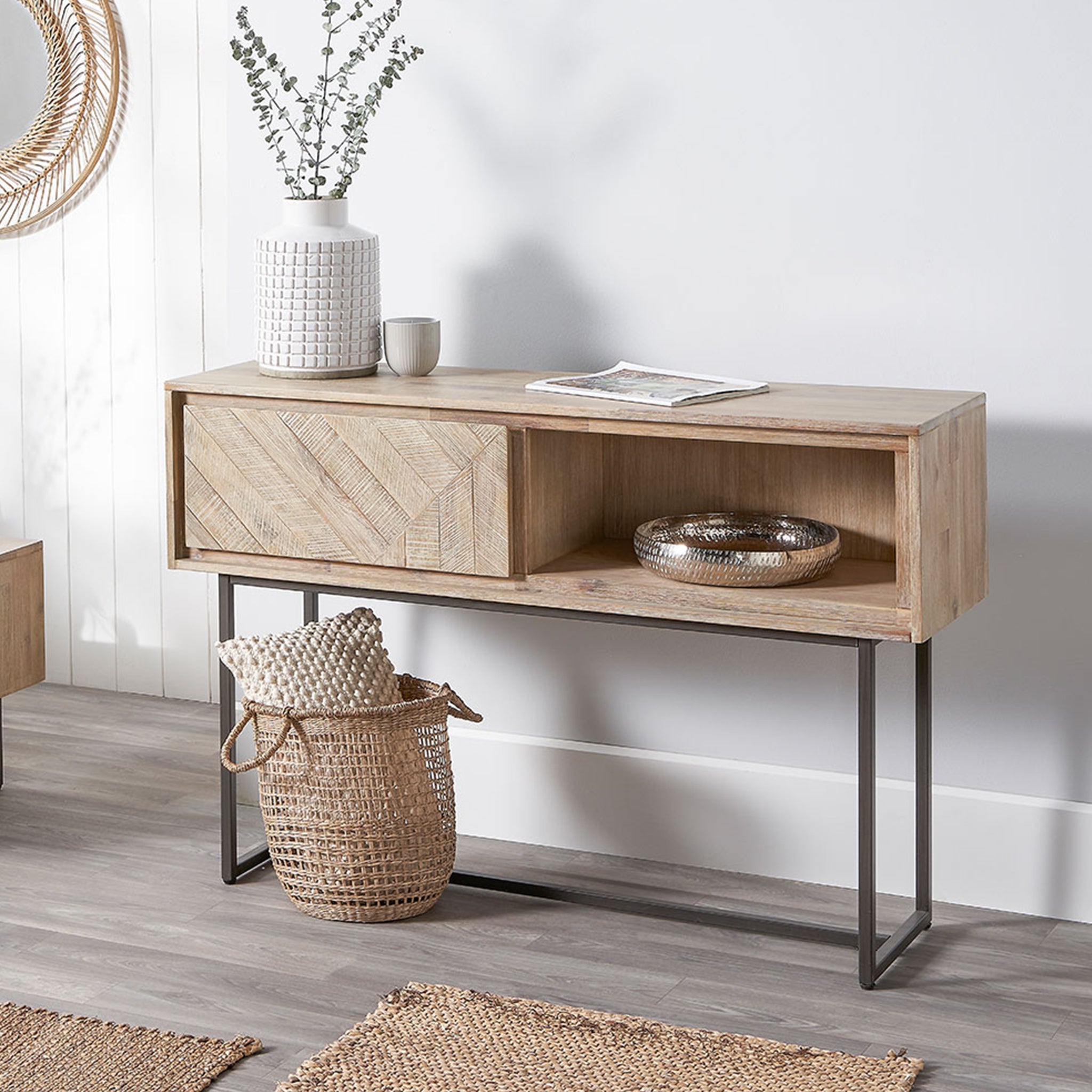 The Pacific Lifestyle - Marca Acacia Wood 1 Drawer Console Table boasts sleek black metal legs. Topped with a vase, candle, and magazine, and complemented by a basket and rug below, it seamlessly enhances any gray floor space with its Scandi style.