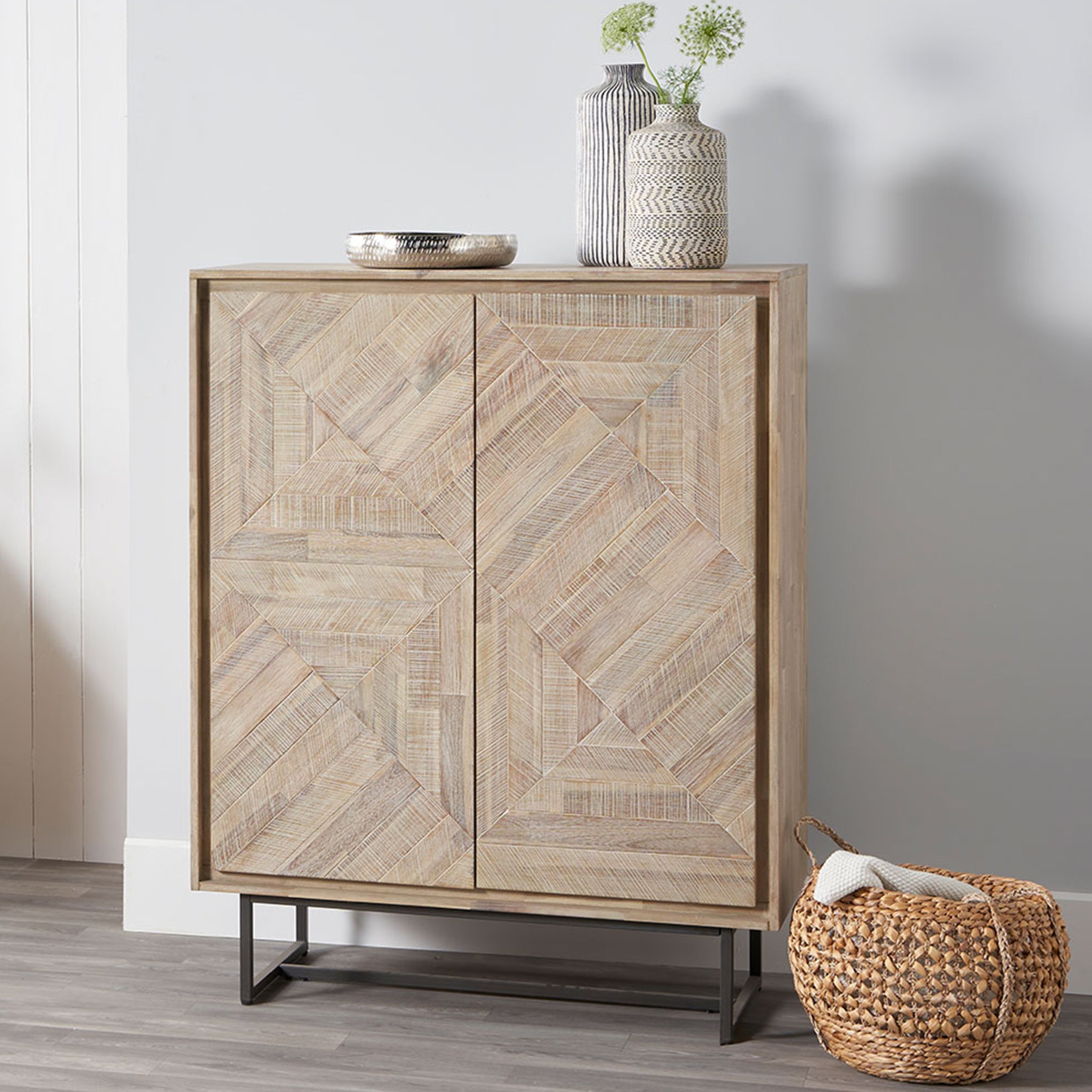 The Pacific Lifestyle - Marca Acacia Wood 2 Door Bar Cabinet features Scandi style, a geometric pattern, and a metal base. Part of the Acacia Collection, it blends form and function with decorative vases and a bowl on top, a basket below, against a light wall, providing ample storage space.